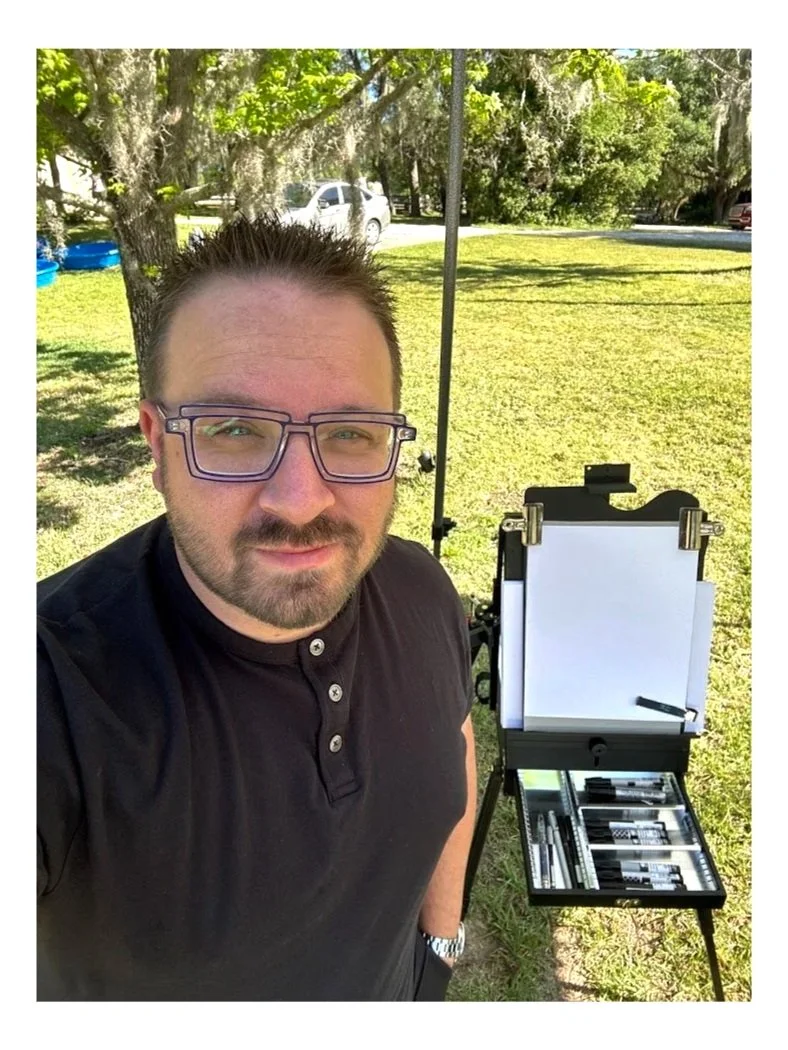 A man with glasses and a beard taking a selfie outdoors on a bright, sunny day. Behind him, there is a large tree with hanging moss, grass, a white car, and a few other vehicles. Next to him, there is a portable case with open compartments containing various tools, possibly precision instruments or small hardware components.