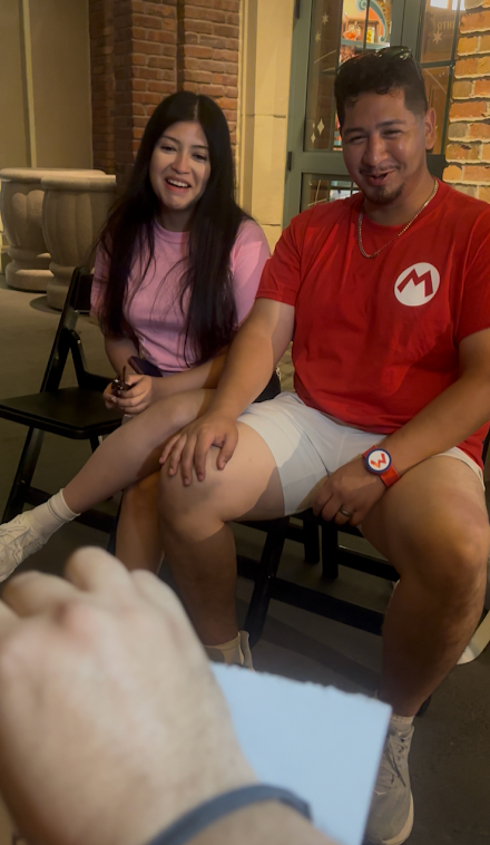 A man and woman sitting on chairs inside a cafe, smiling and relaxing. The man is wearing a red Mario shirt, white shorts, and a smartwatch. The woman is wearing a pink shirt and white sneakers. In the foreground, a dog is partially visible, held by someone outside the frame.