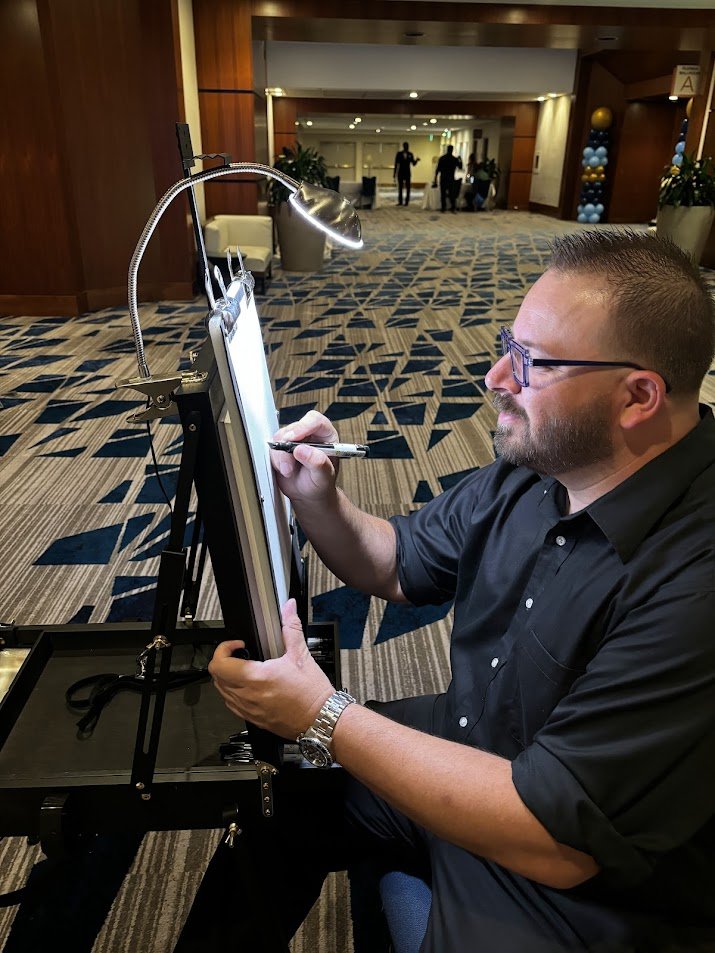 A man with glasses and a beard, wearing a black shirt, sits at a table and writes on a whiteboard. The setting appears to be a hotel lobby or conference area with patterned carpet, chairs, and balloons in the background.