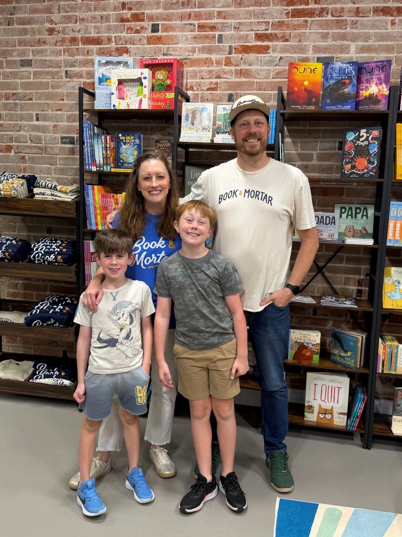 A group of four people, two adults and two children, posing together inside a bookstore with bookshelves filled with colorful children's books and graphic novels behind them. The adults are smiling and wearing casual clothes, while the children are standing in front of them.