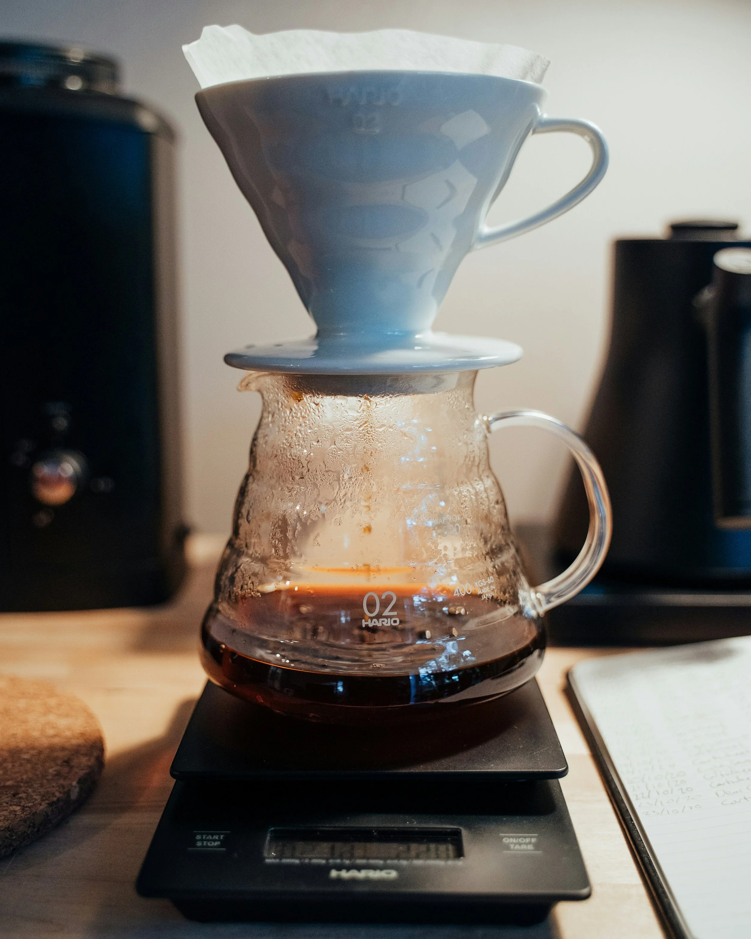 A pour-over coffee maker with a paper filter on top, placed on an electronic scale that is turned on, with a glass carafe underneath collecting dark brewed coffee. There are kitchen appliances visible in the background, and a handwritten note is seen on the right side of the image.