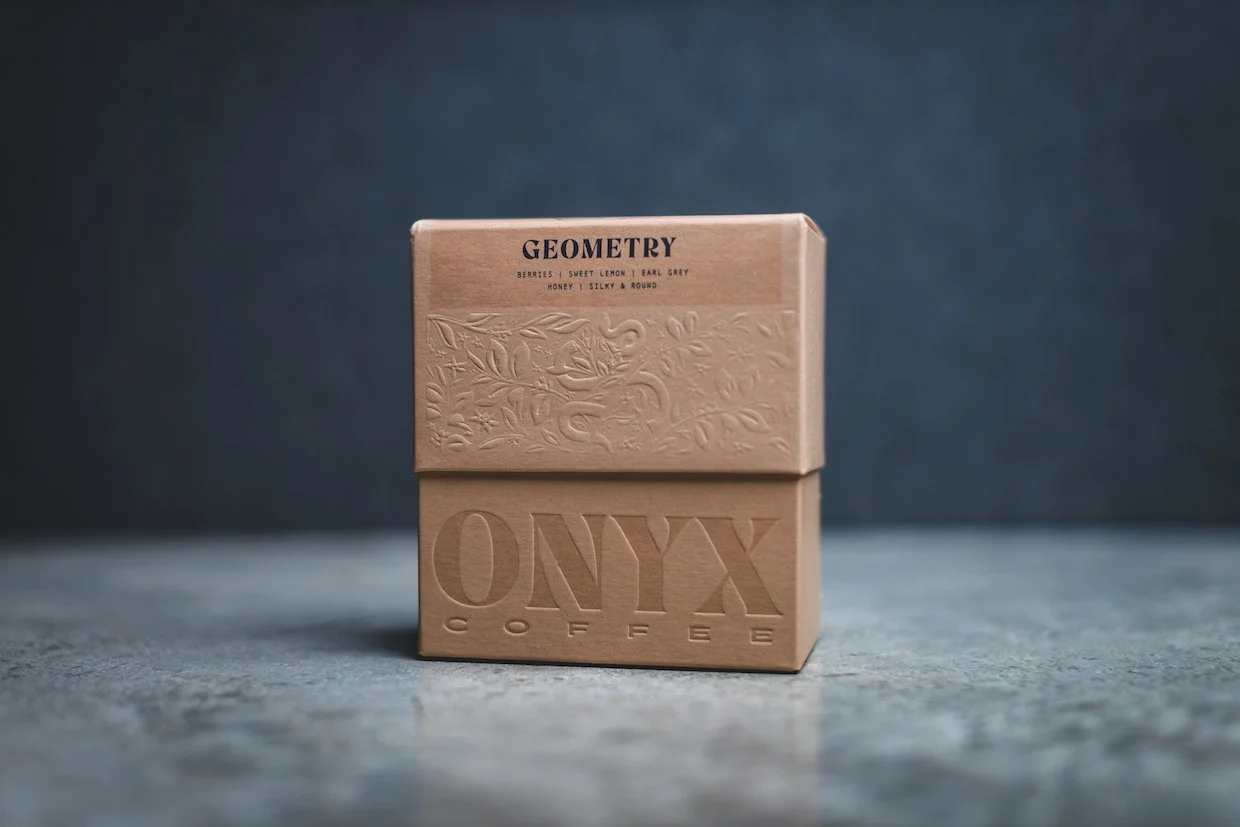 A beige box with the brand name 'ONYX Coffee' embossed on the front, placed on a surface with a dark background. The top section of the box has a textured design and the word 'GEOMETRY' along with a flavor description.