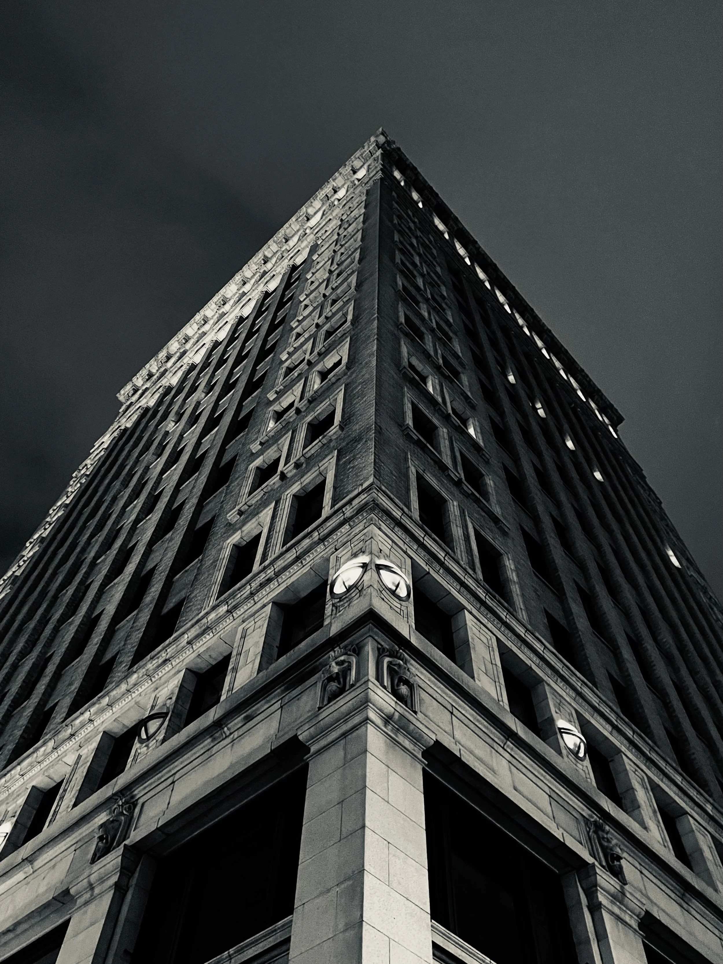 Looking straight up at the sharp corner of a dark ornate high-rise, its decorative stonework receding into a near-black sky. Two globe lights glow at the building’s base, small against the mass above.