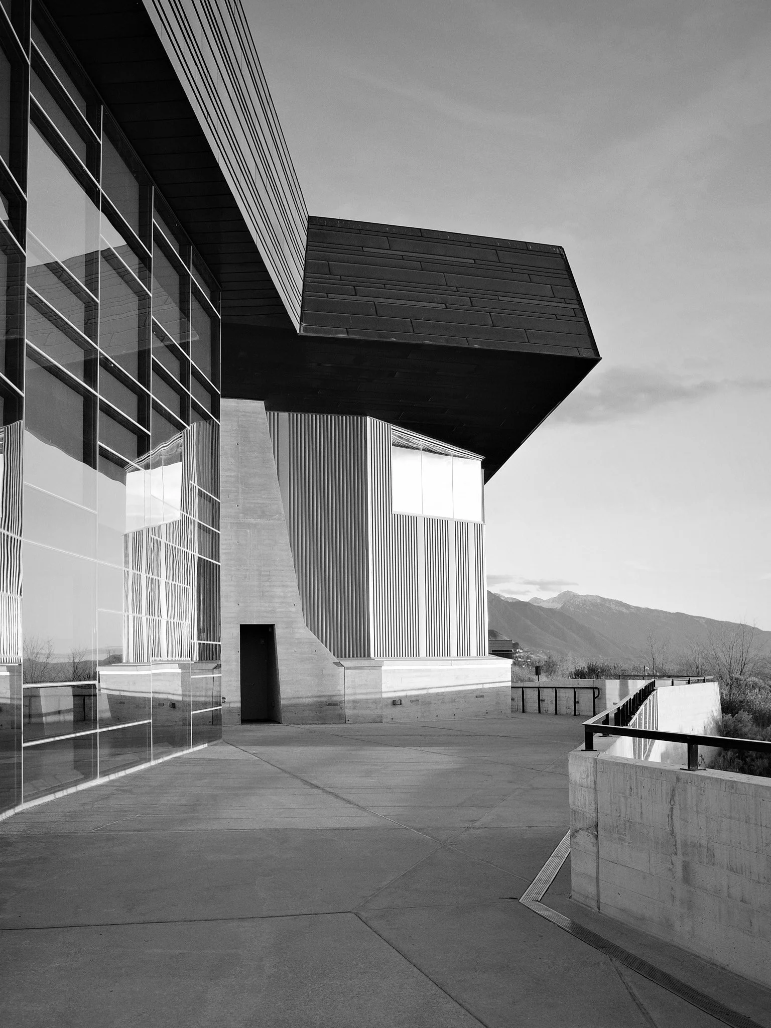 The cantilevered roof of a contemporary building juts into a pale sky, glass walls reflecting the surrounding landscape. The Wasatch Mountains rise in the distance, framed between architectural forms.