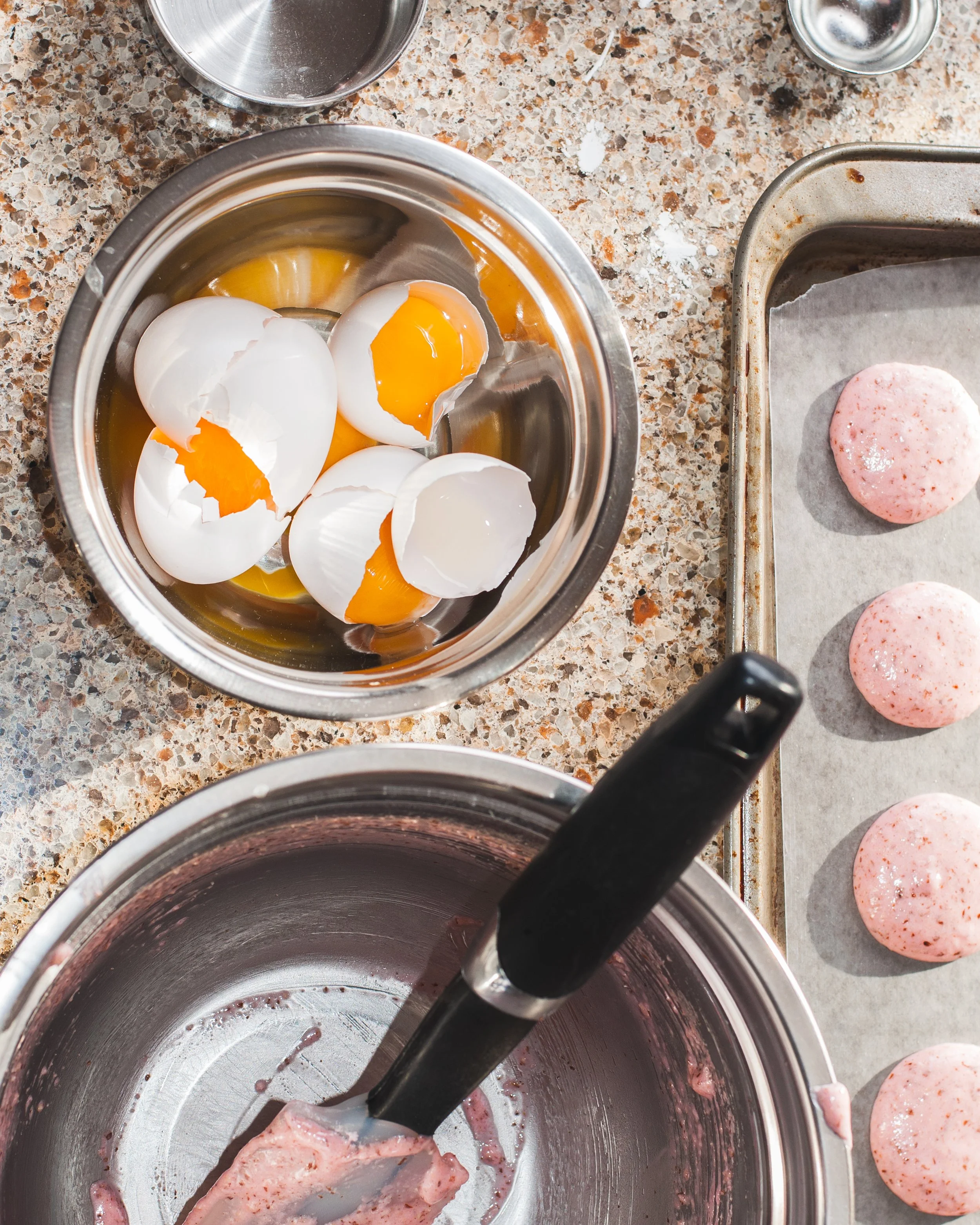 Baking flat lay with cracked eggs mixing bowl and pink macaron shells on marble surface overhead food photography