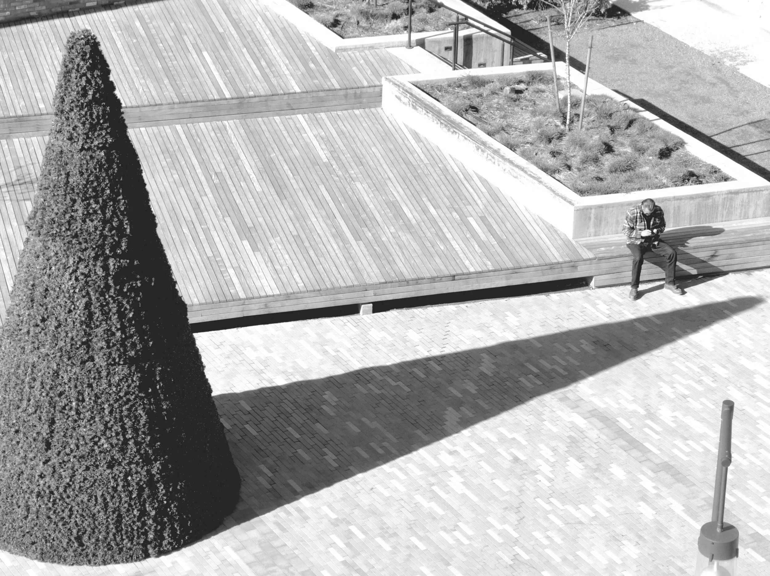 An overhead view of a geometric urban plaza — wood decking, brick pavers, a cone-shaped topiary casting a long shadow. A single person sits at the edge of the platform, looking down at their phone.