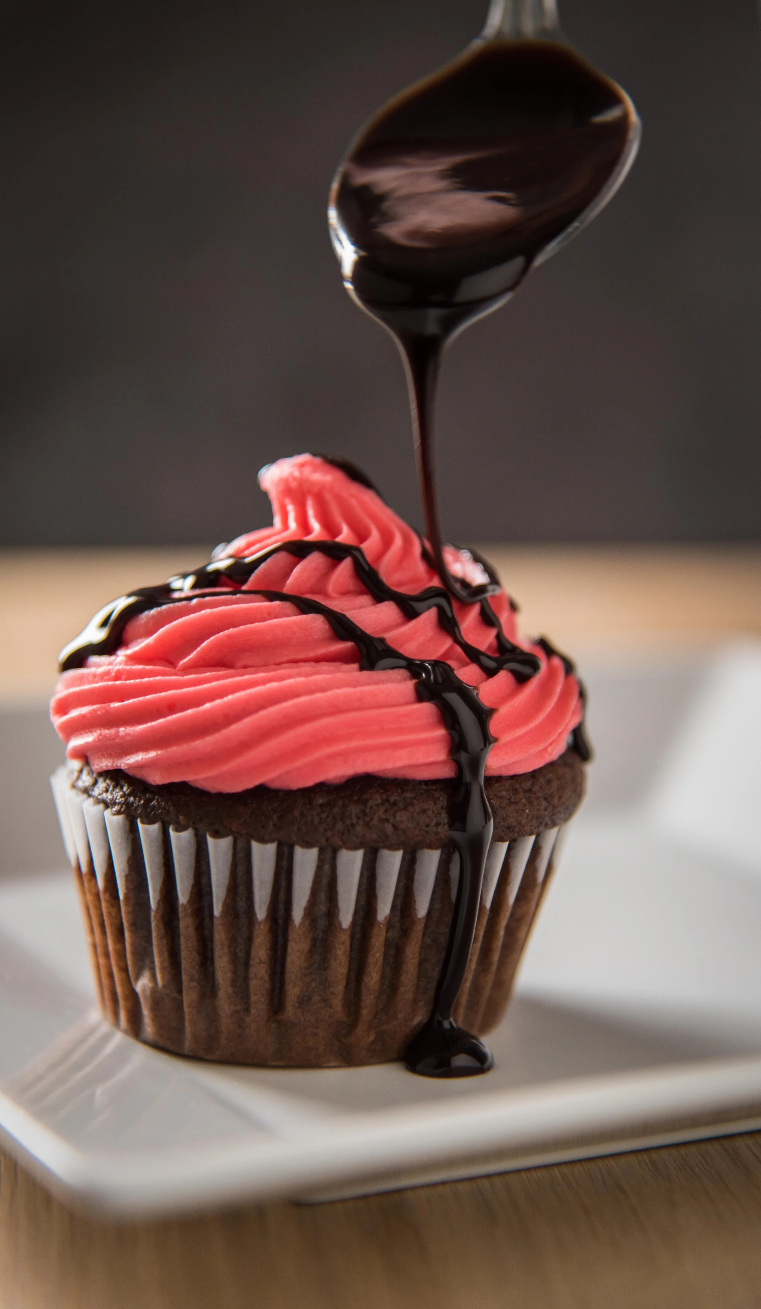 Chocolate cupcake with pink frosting and chocolate sauce dripping from spoon on white plate dramatic food photography salt lake city