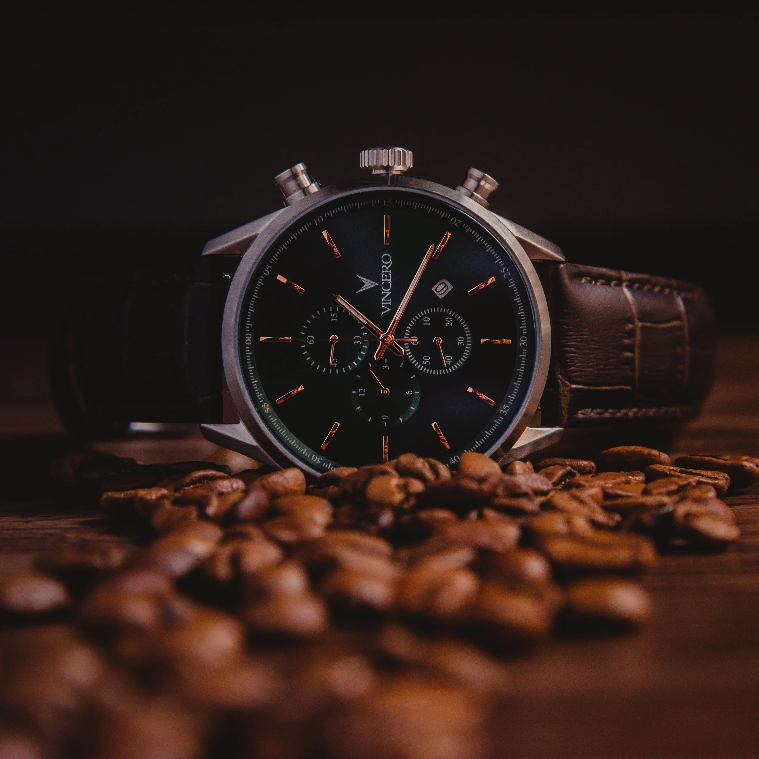 Vincero chronograph watch surrounded by coffee beans on dark wooden surface dramatic product photography