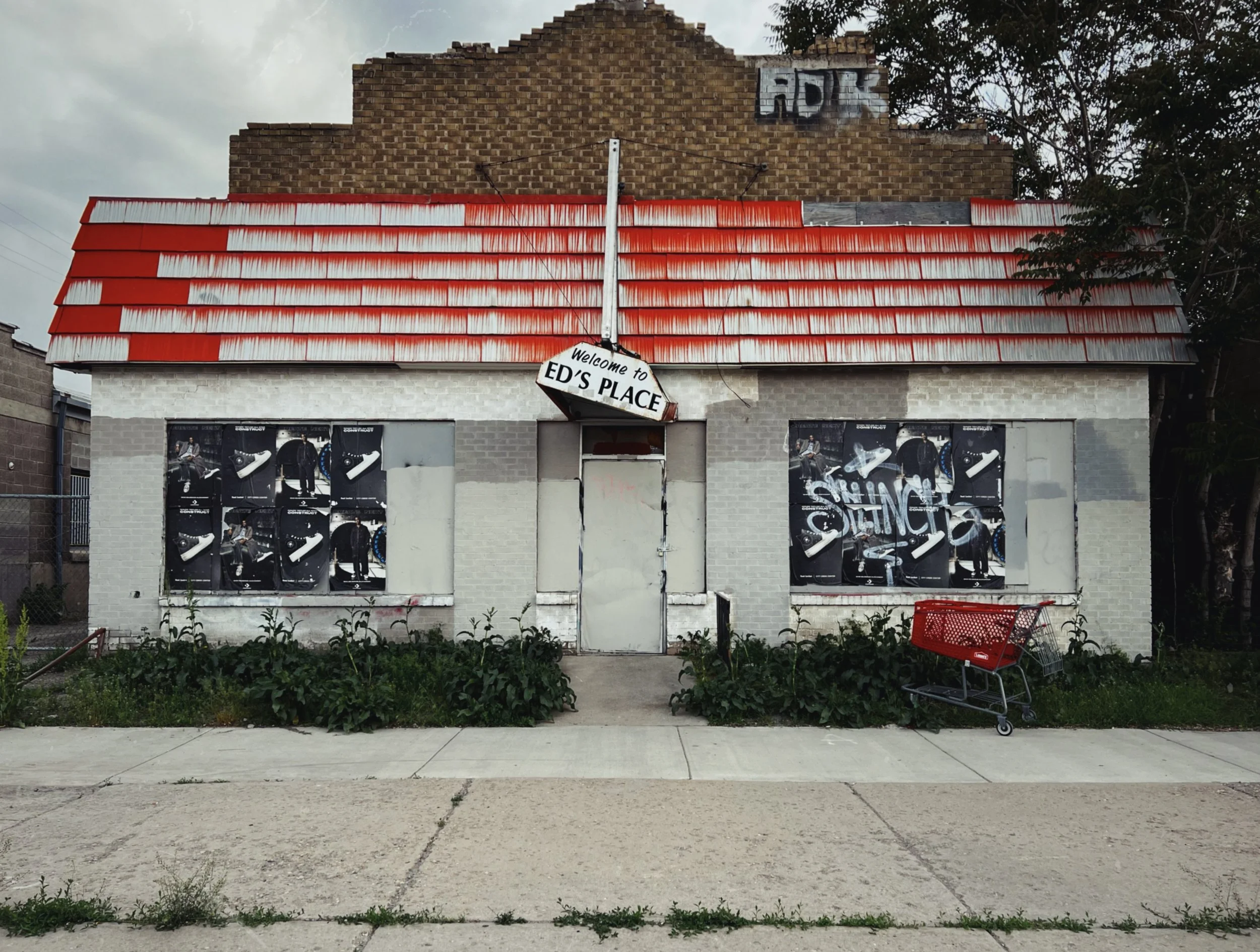 A shuttered storefront with a tilted Welcome to Ed’s Place sign, red-fringed awning peeling, windows papered over with old posters. A lone red shopping cart sits in the overgrown lot beside it.