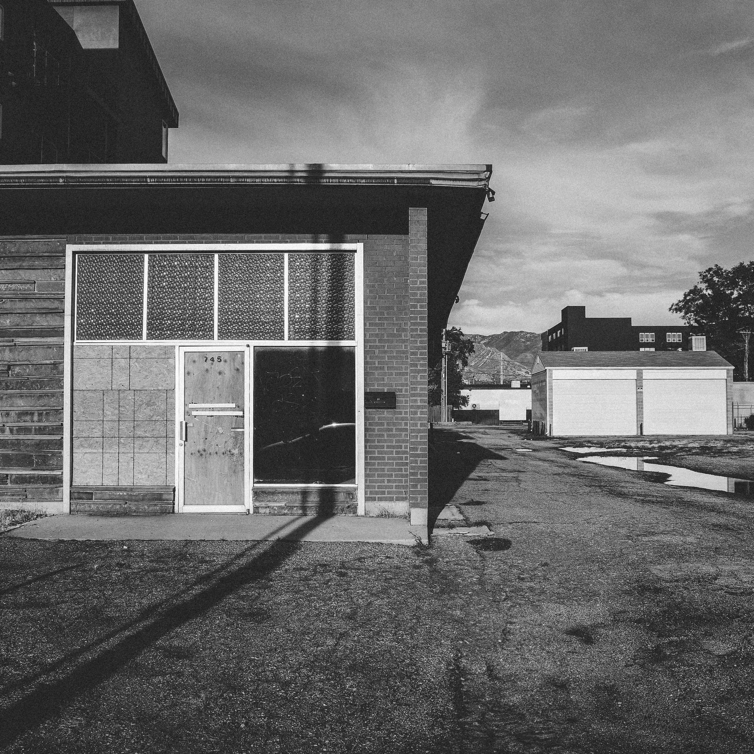 A boarded commercial building at address 745, its long shadow stretching across an empty gravel lot. Mountains sit quietly in the distance under a dramatic cloud-streaked sky.”
