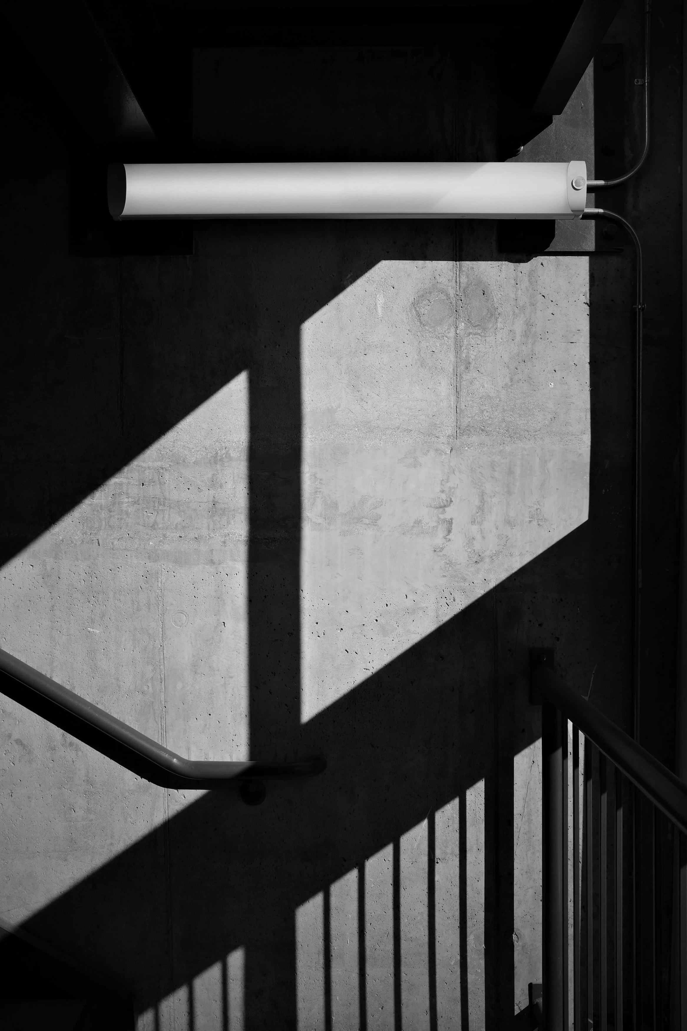 A white cylindrical light fixture mounted on a concrete wall, casting a sharp angular shadow that cuts the frame into geometric planes of light and dark. A railing repeats the pattern below.