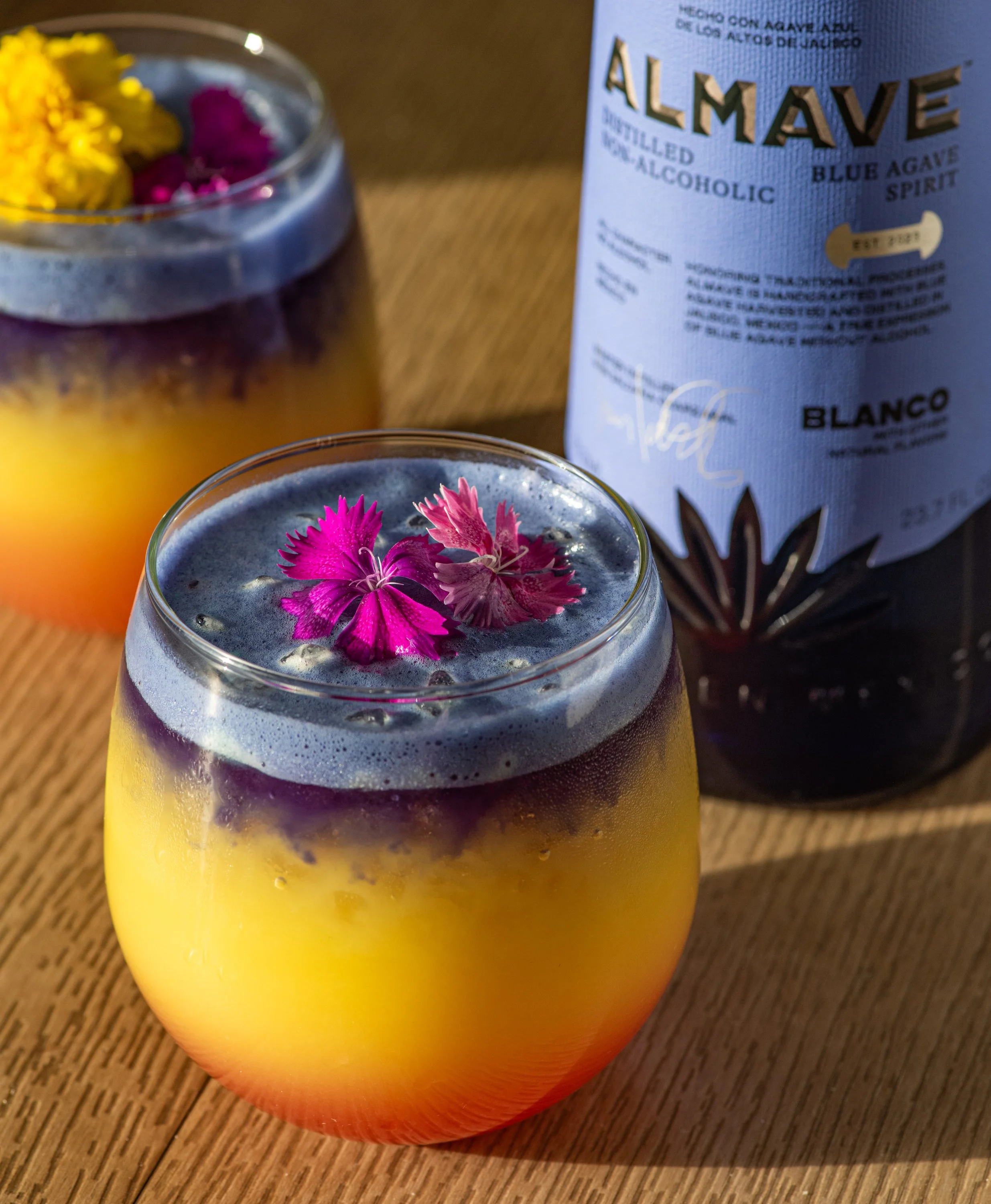 Almave blue agave spirit nonalcoholic cocktail with layered sunset colors and pink flower garnish on wooden surface beverage photography Salt Lake City