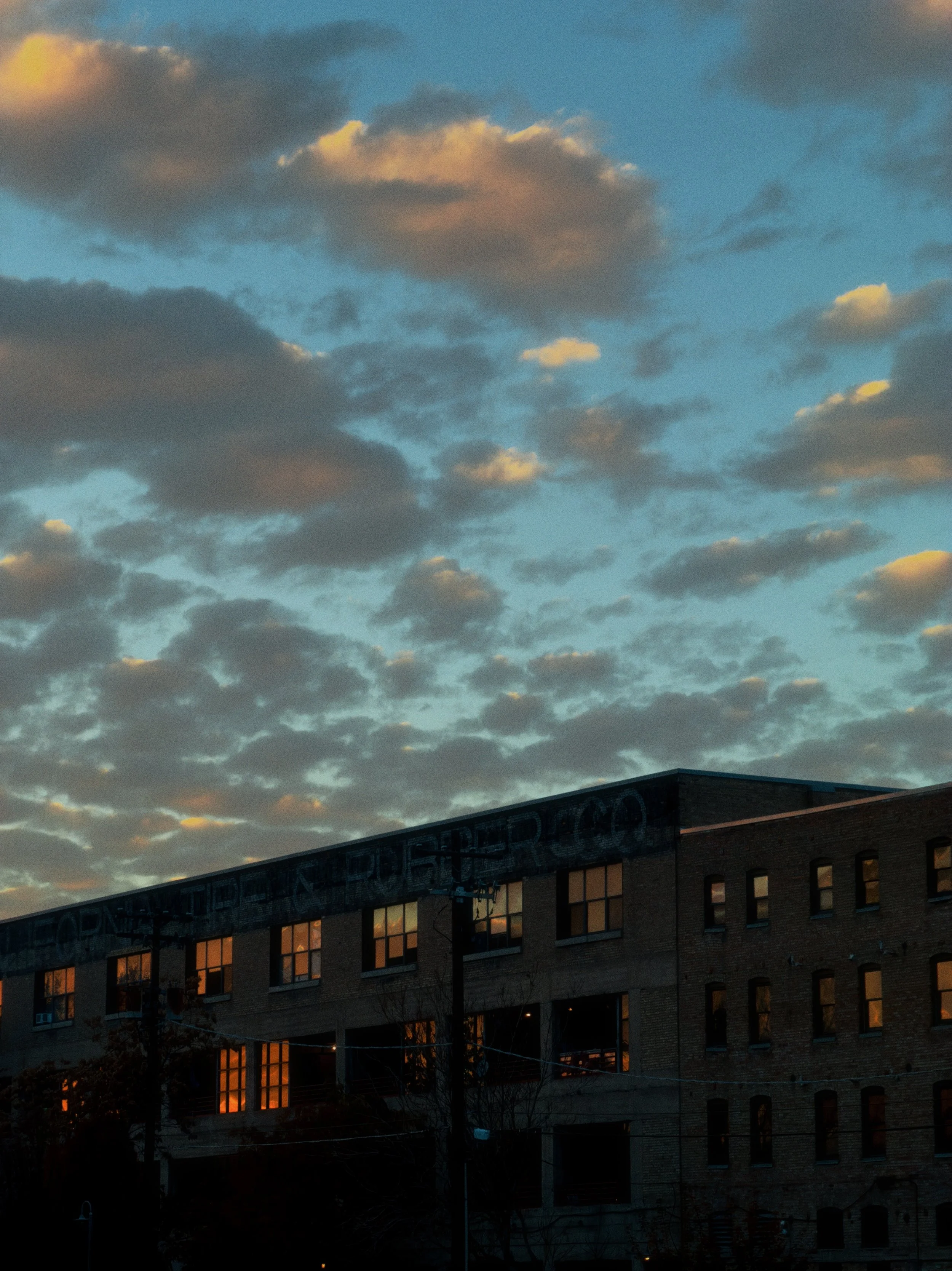 A long brick warehouse at blue hour, its windows glowing amber. Graffiti runs along the roofline. Gold-edged clouds drift above in a deepening blue sky.
