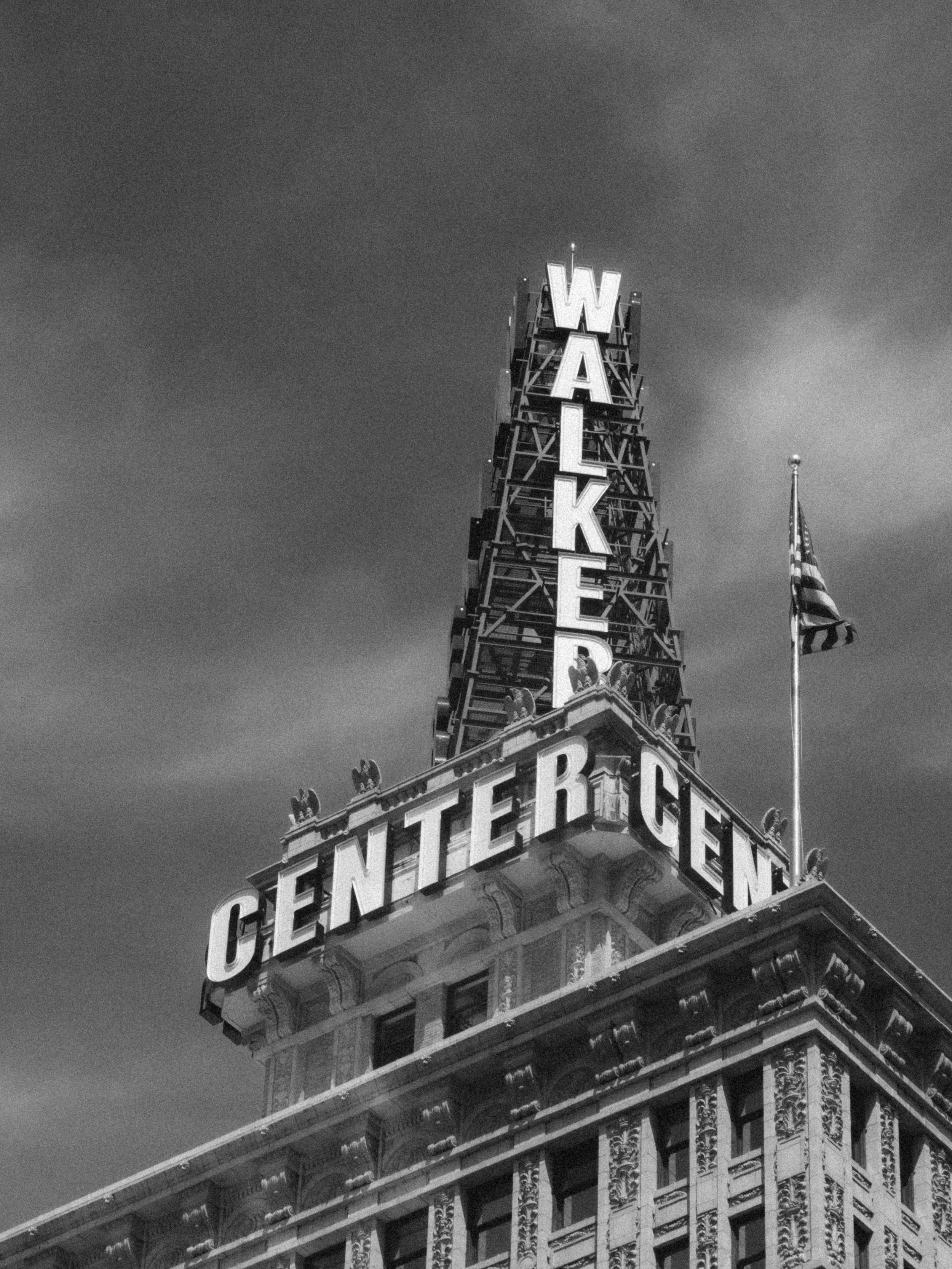 The pyramidal sign tower of Salt Lake City’s Walker Center building rises against a stormy black-and-white sky, the letters W-A-L-K-E-R stacked vertically in illuminated panels. An American flag catches wind to the right.