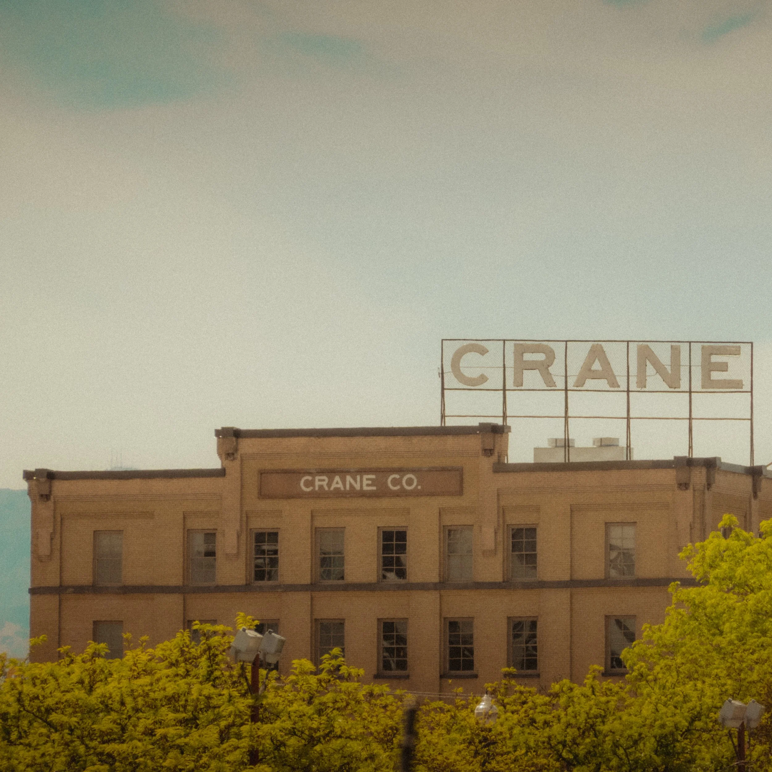 The weathered facade of the Crane Co. building in Salt Lake City, shot through a veil of spring trees. A large rooftop sign spells CRANE against a pale, hazy sky in warm filmic tones.