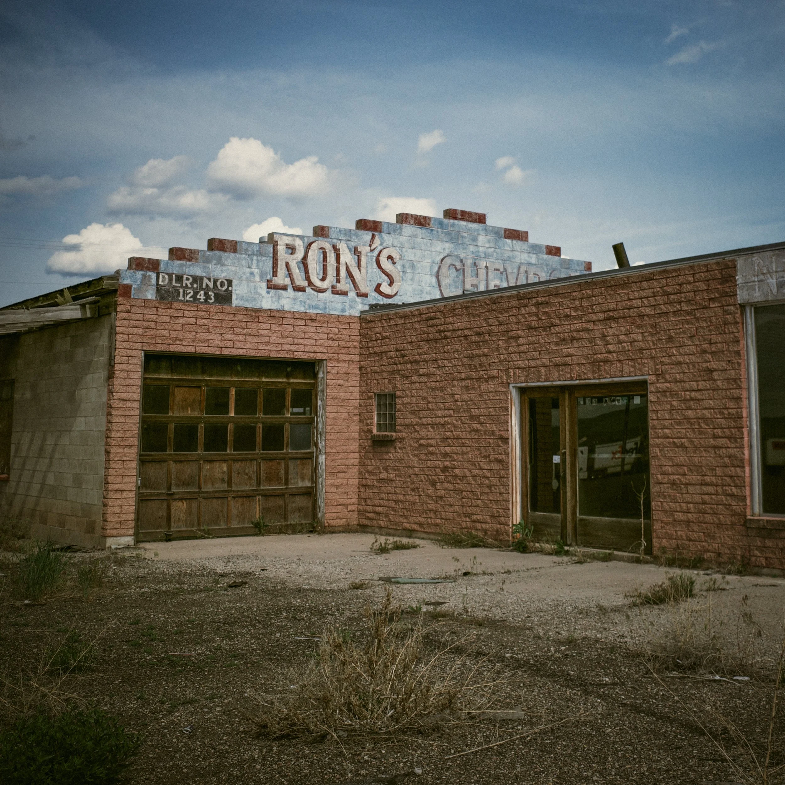 An abandoned brick dealership in Beaver, Utah, its painted Ron’s Chevrolet sign fading above boarded garage doors. Weeds push through cracked pavement. The sky is wide and indifferent.