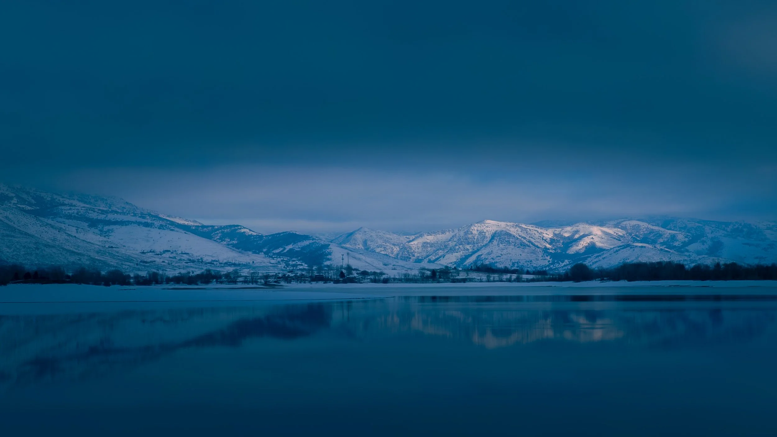 Snow-covered mountains reflected in a still lake at blue hour, the landscape bathed in deep indigo light beneath a stormy winter sky.