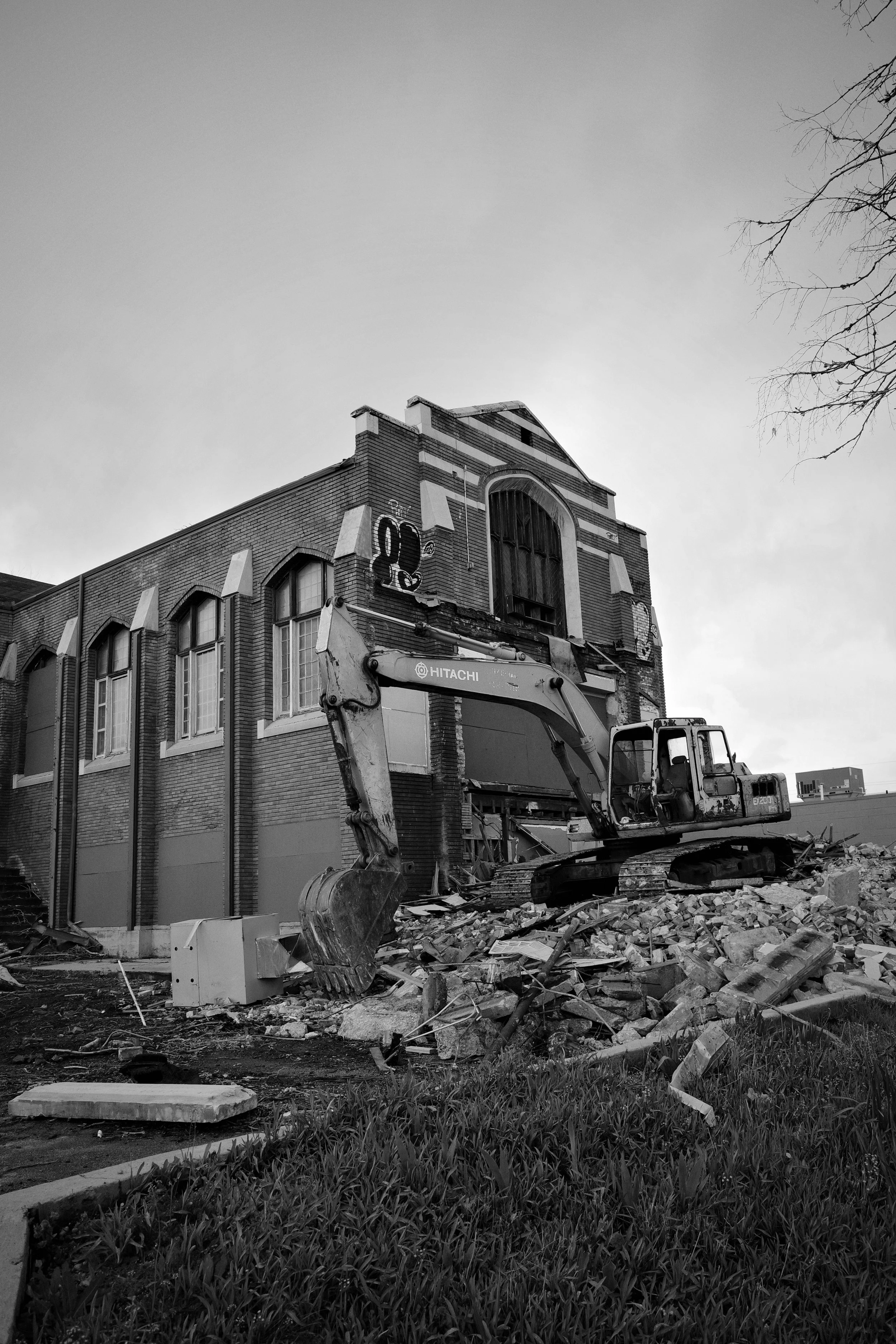 A Hitachi excavator tears into the facade of an ornate brick building, rubble scattered across the foreground. The building’s arched windows and decorative brickwork remain intact above the destruction.
