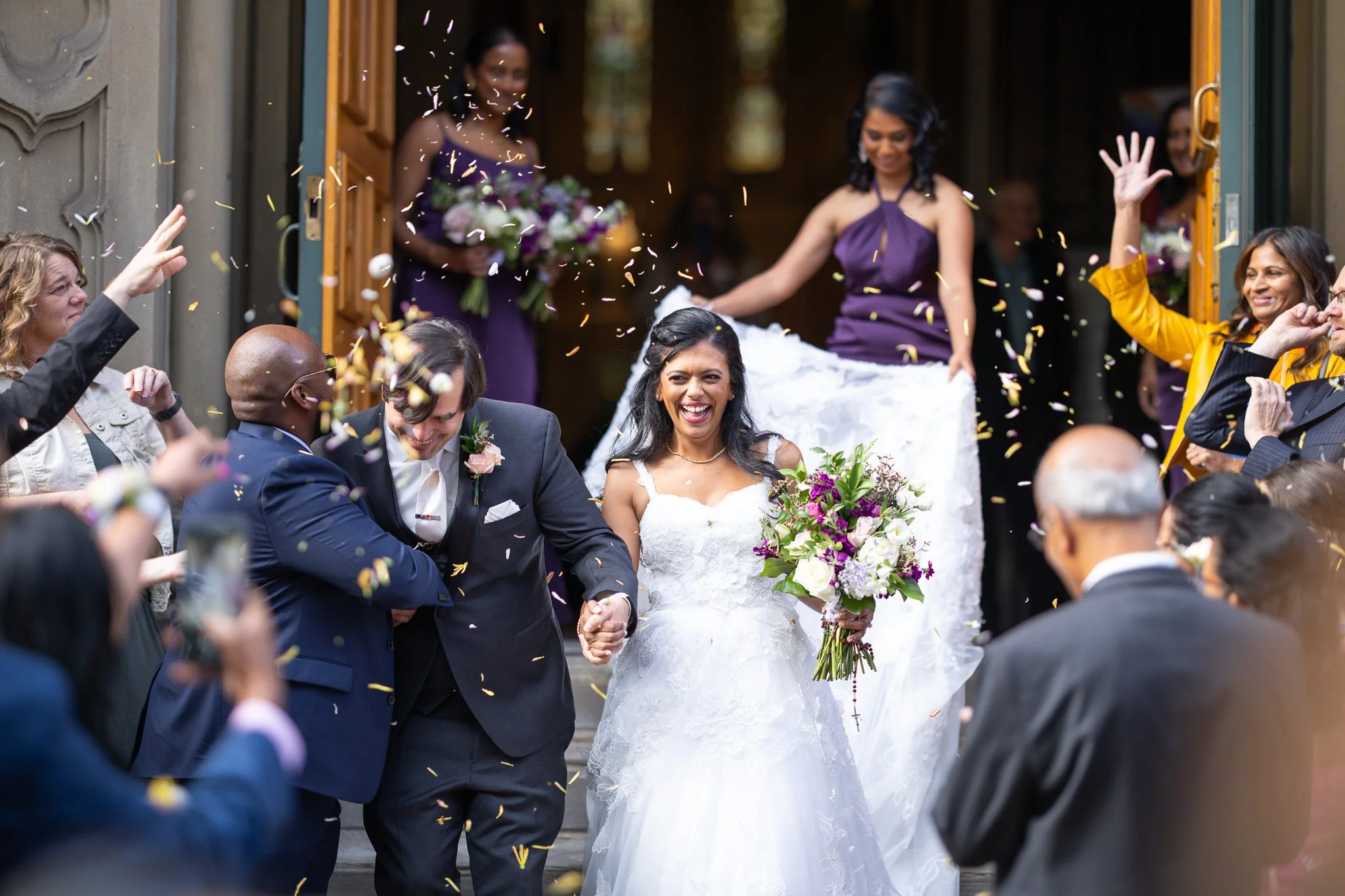 A bride and groom holding hands, smiling, as guests celebrate with confetti, outside a building.