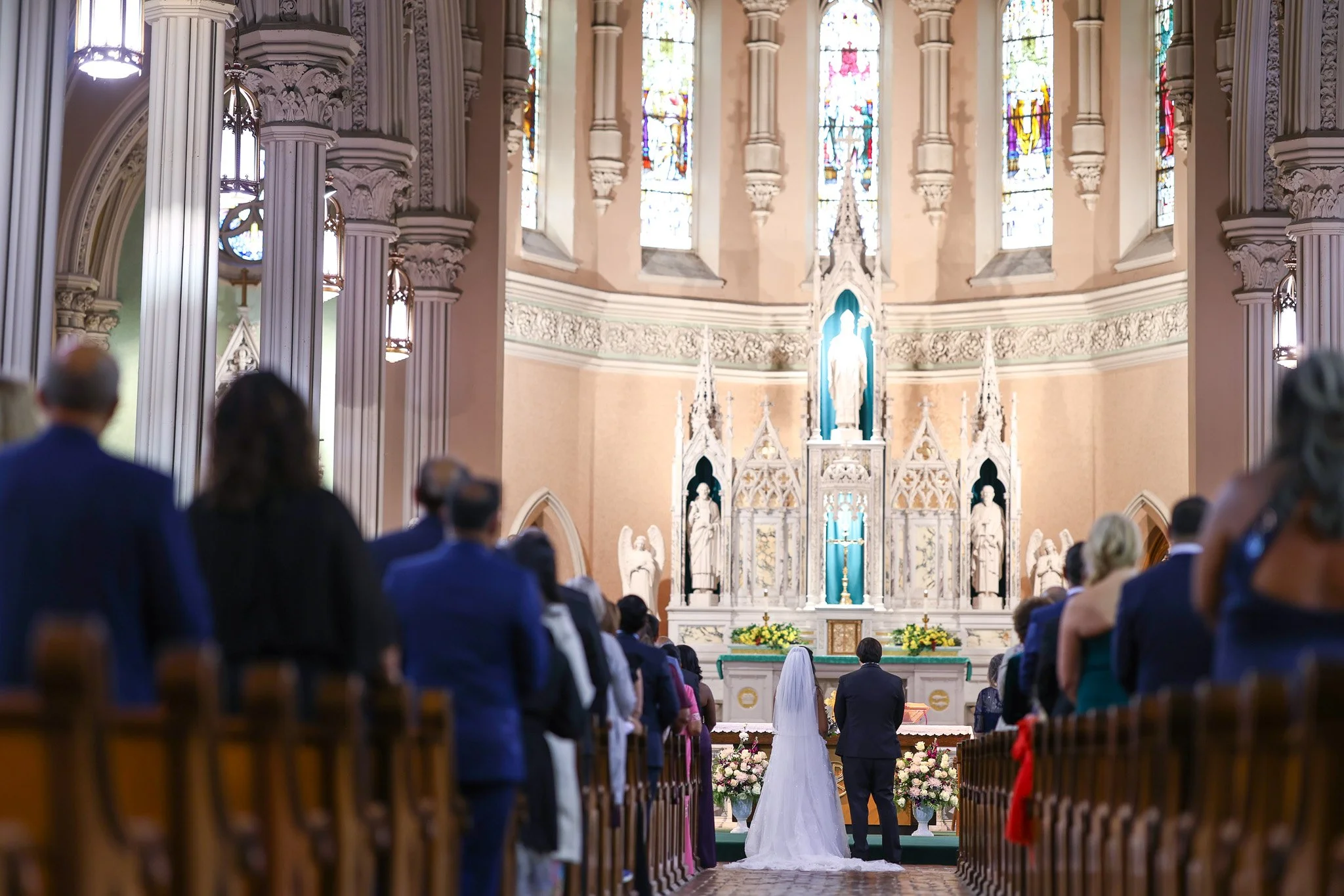 A wedding ceremony taking place inside a grand church with stained glass windows and ornate architecture. The bride and groom are standing at the altar facing the priest, with guests seated in pews observing.