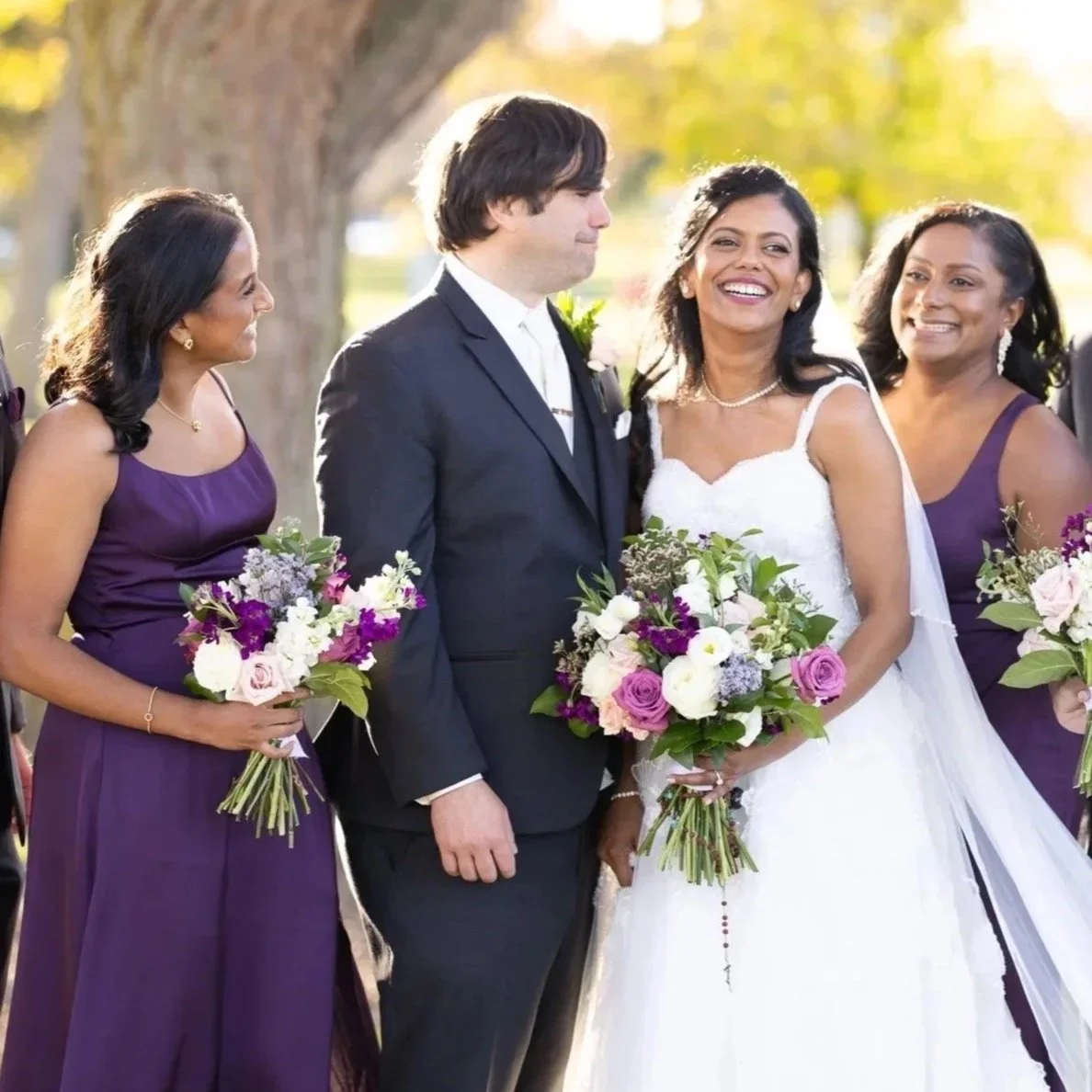 Wedding scene with a bride in a white gown and groom in a dark suit, surrounded by bridesmaids in purple dresses holding bouquets, outdoors during daytime.