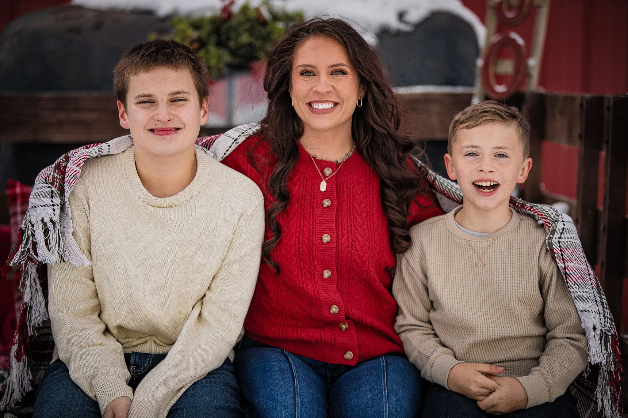 A smiling woman with long dark hair and two young boys sitting close together outdoors, wearing warm sweaters, with a wooden fence and red barn in the background.