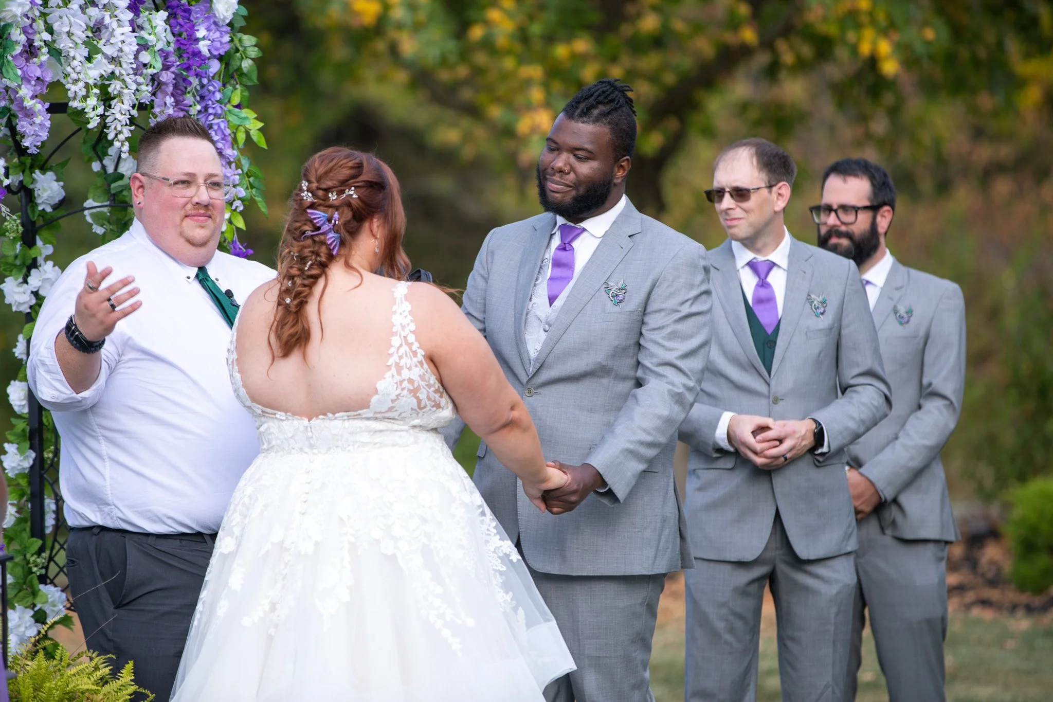 A wedding ceremony outdoors with five people, including the bride and groom holding hands, with a floral arch in the background and a blurred natural setting.