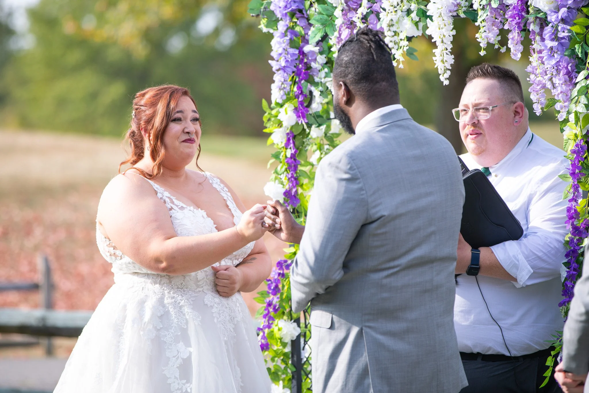 A woman with red hair in a white wedding dress holding hands with a man in a light gray suit during a wedding ceremony outdoors, with another man in a white shirt and glasses standing nearby. They are under a floral arch with purple, white, and green