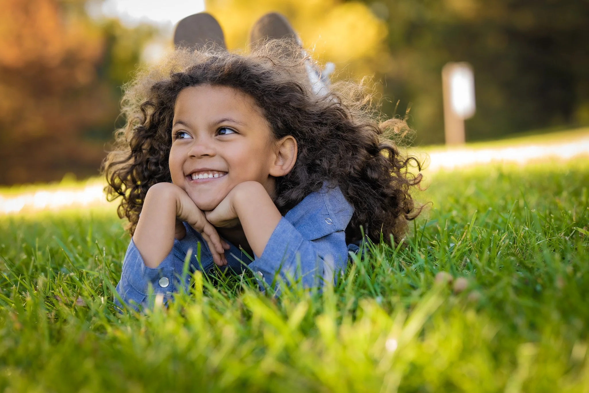 A young girl with curly hair lying on grass outdoors in a park, smiling and resting her chin on her hands, with trees and a signpost in the background during autumn.
