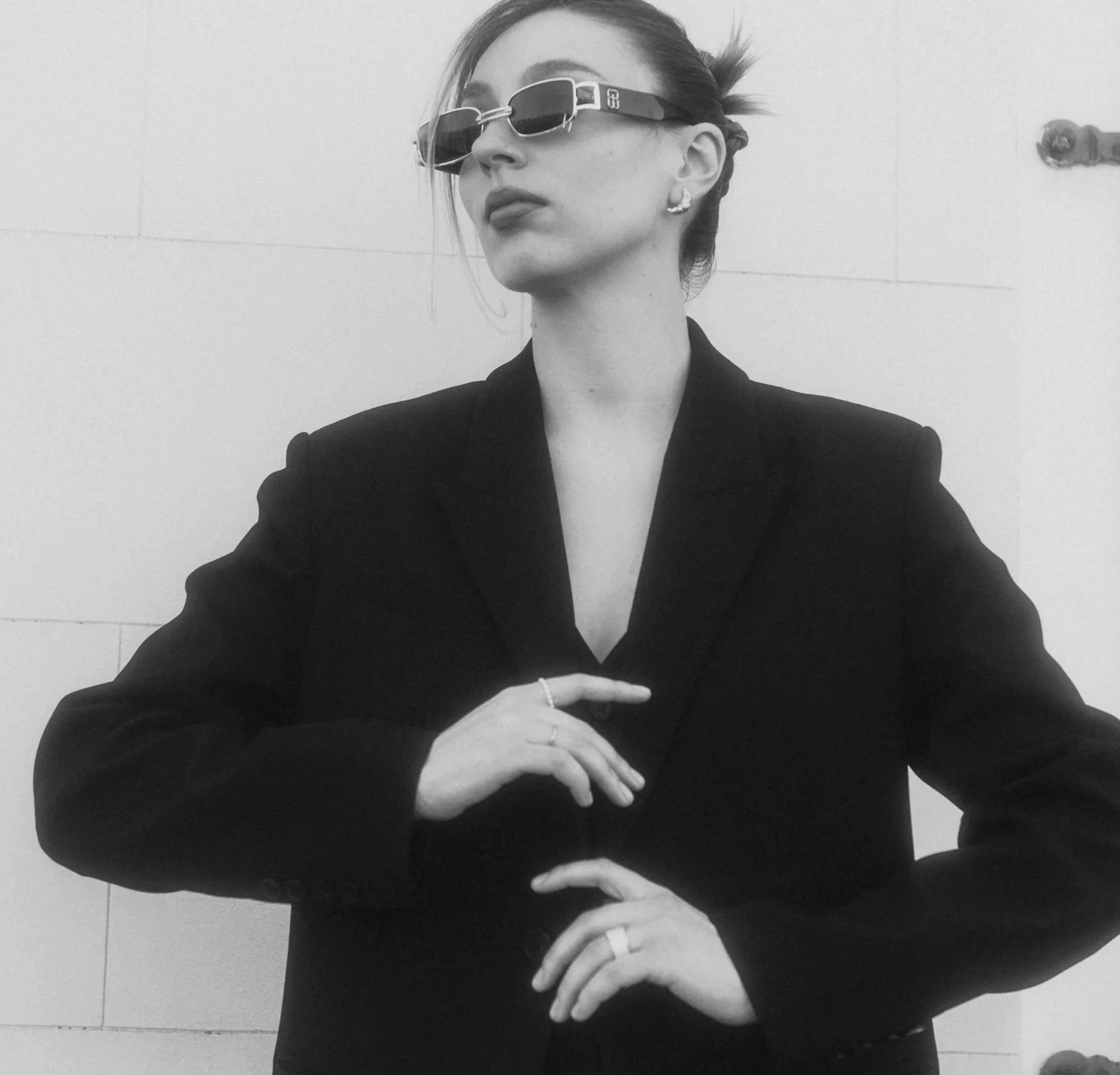Black and white photo of a woman wearing sunglasses and jewelry, with her hand on her chest. She is dressed in a dark blazer with a deep V-neck. Her hair is styled back, and she is looking at an angle.