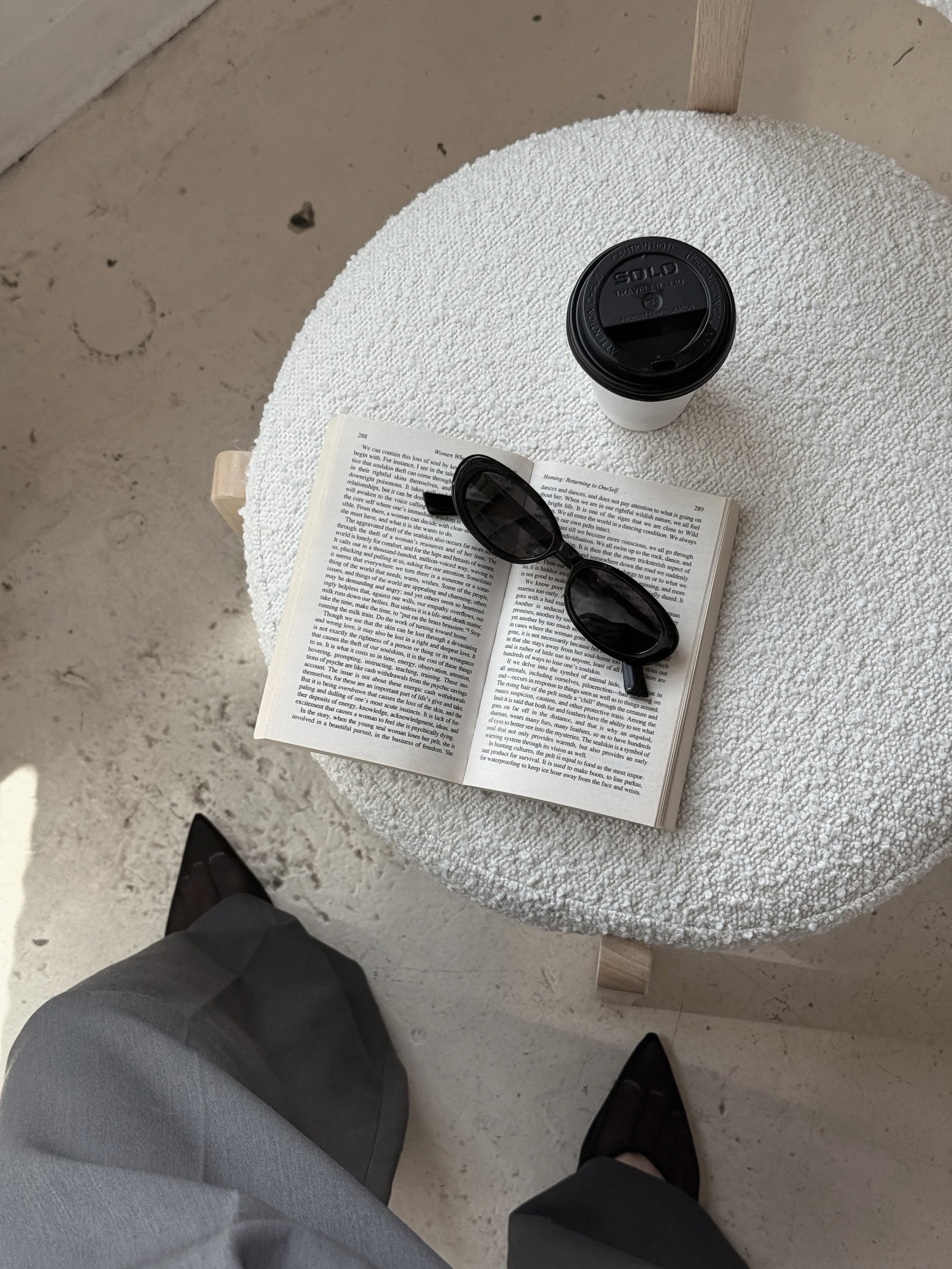 A white round table with a textured surface, a cup of coffee with a black lid, an open book, black sunglasses, and a person dressed in gray pants and black shoes standing nearby.
