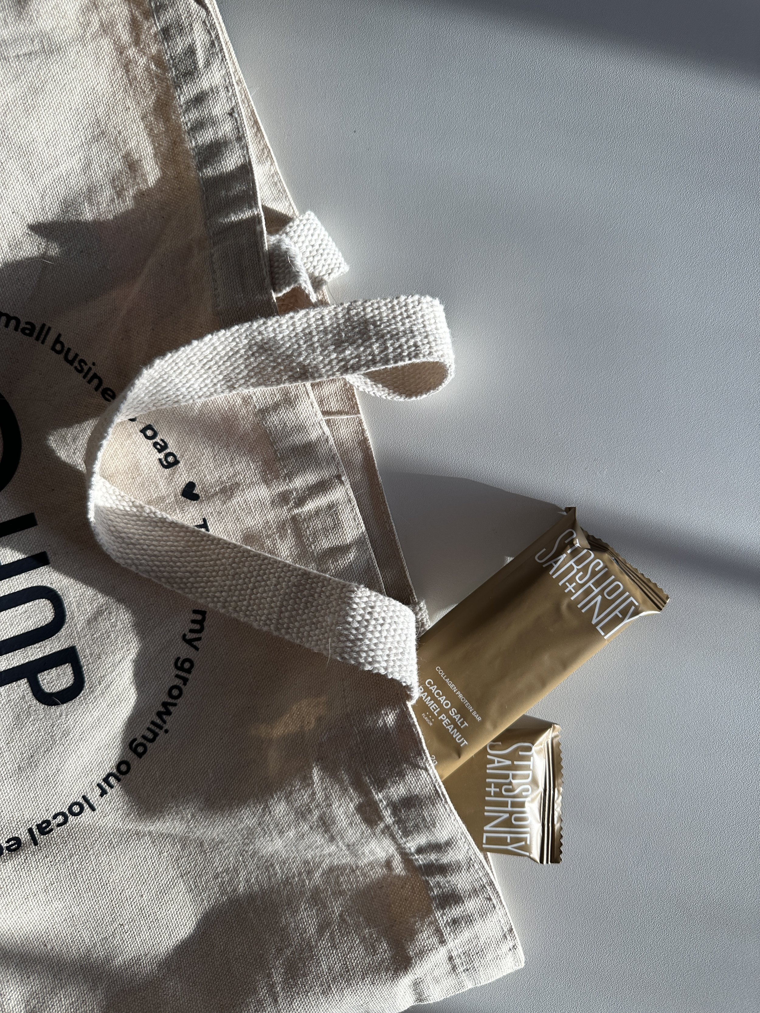 Close-up of a beige canvas tote bag with black text, a white fabric handle, and a gold-colored snack package labeled 'SOURCES JAR + HEALTHY'.