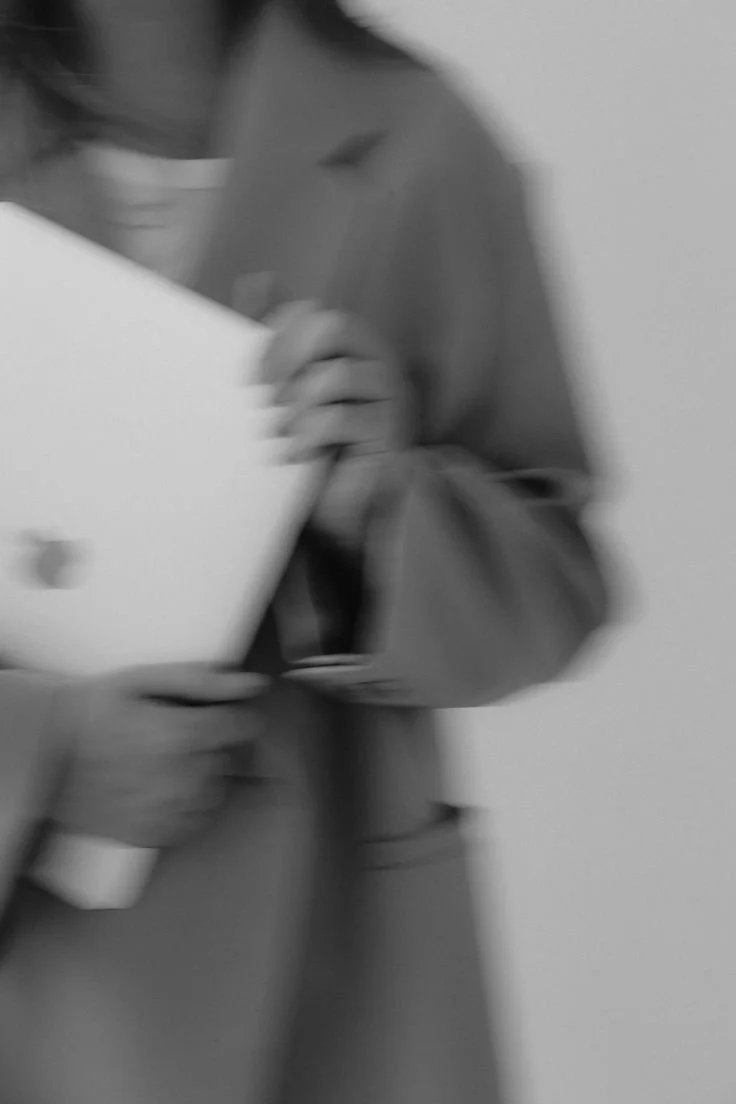 A person in a trench coat holding an Apple laptop, with the image in black and white and slightly blurry.