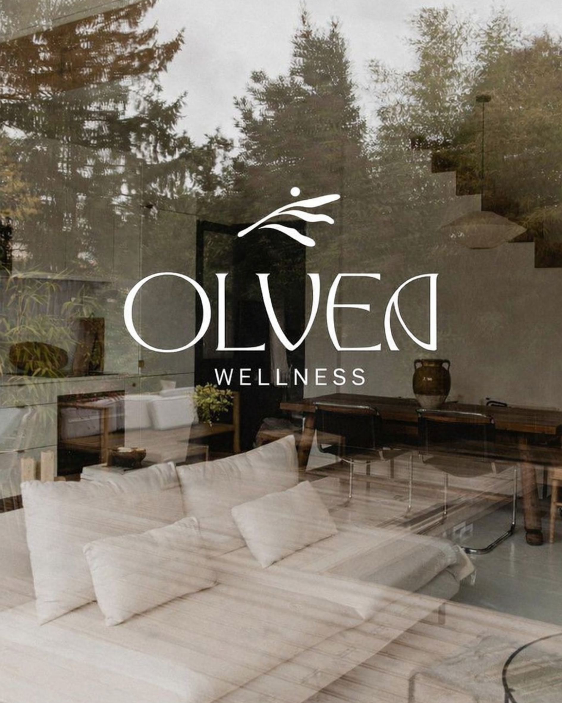 Interior of a wellness or spa center with white couches, a wooden table, plants, and decorative vases, seen through a glass door with a wellness logo.