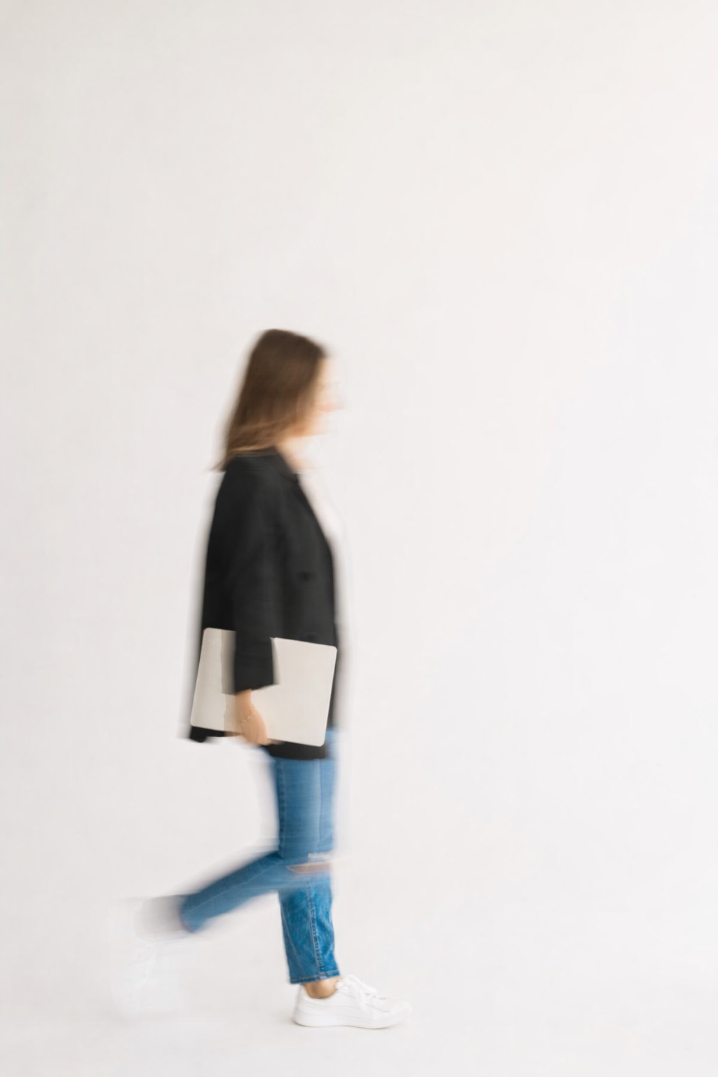 Blurry image of a woman walking while carrying a laptop against a plain white wall.