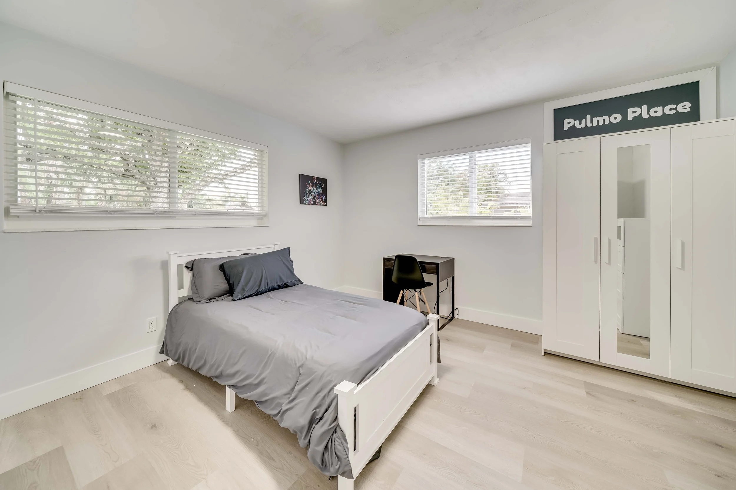 A bedroom with a white bed frame and gray bedding, two windows with blinds, a small black desk with a chair, a framed colorful artwork, and a white wardrobe with a sign that says 'Pulmo Place'.