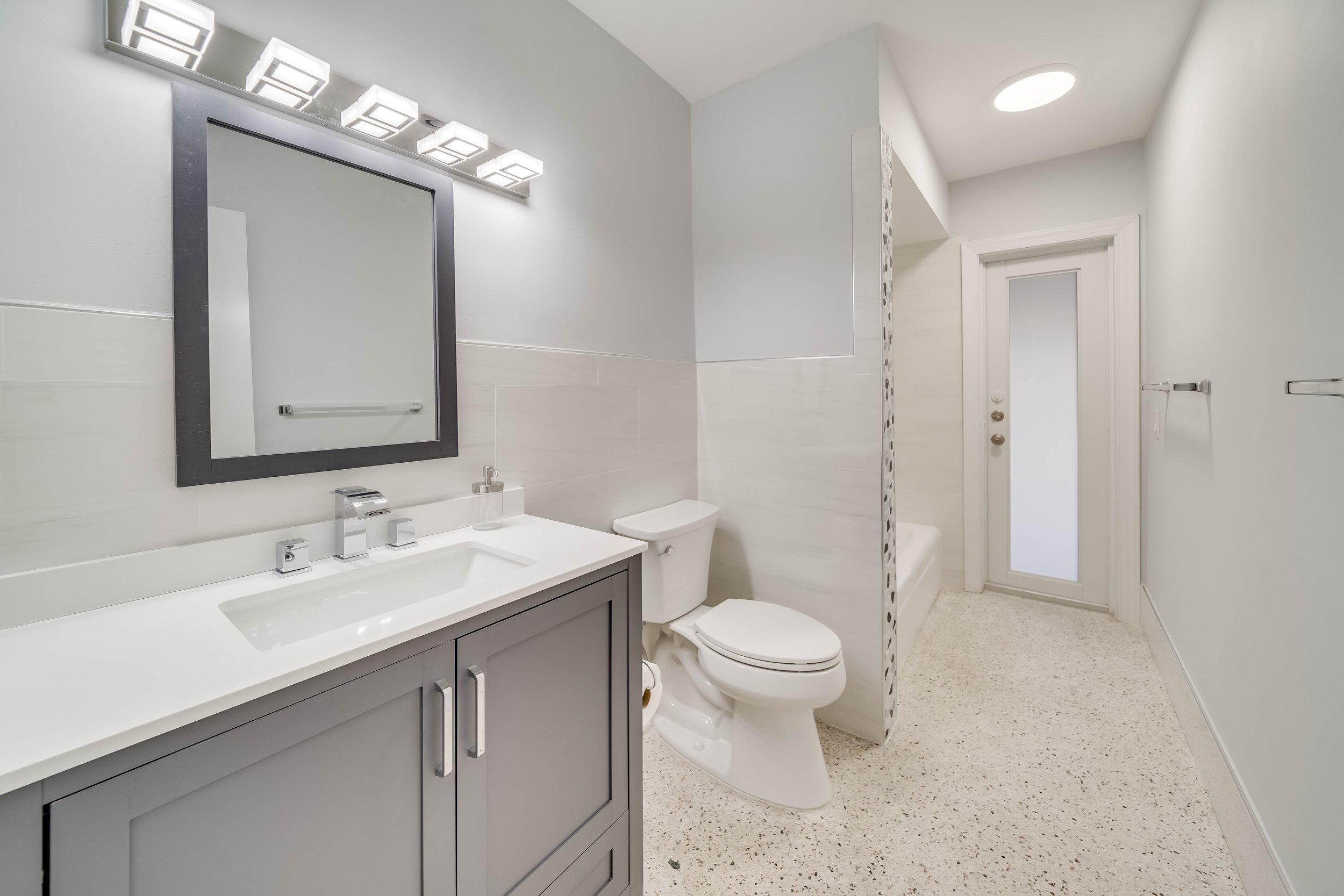 A modern bathroom with a white sink and vanity, a large mirror with square light fixtures, a toilet, and a partial view of a shower with a glass door, white walls, and a small frosted window.