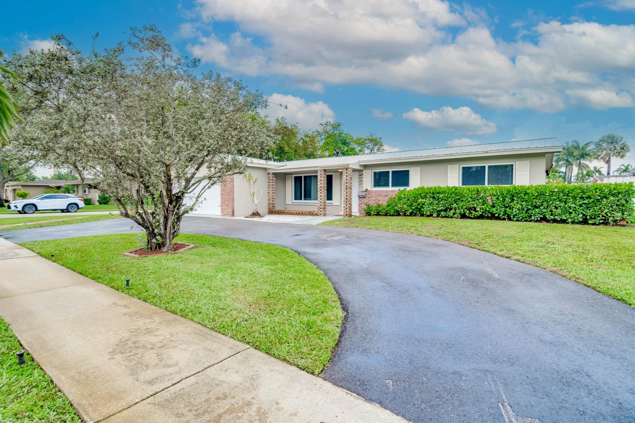 A suburban house with a white exterior, brick accents, and a front yard with green grass and shrubbery. There is a driveway leading to a garage and a silver car parked on the street. A large tree with blooming flowers is in the front yard. The sky is partly cloudy with blue patches.