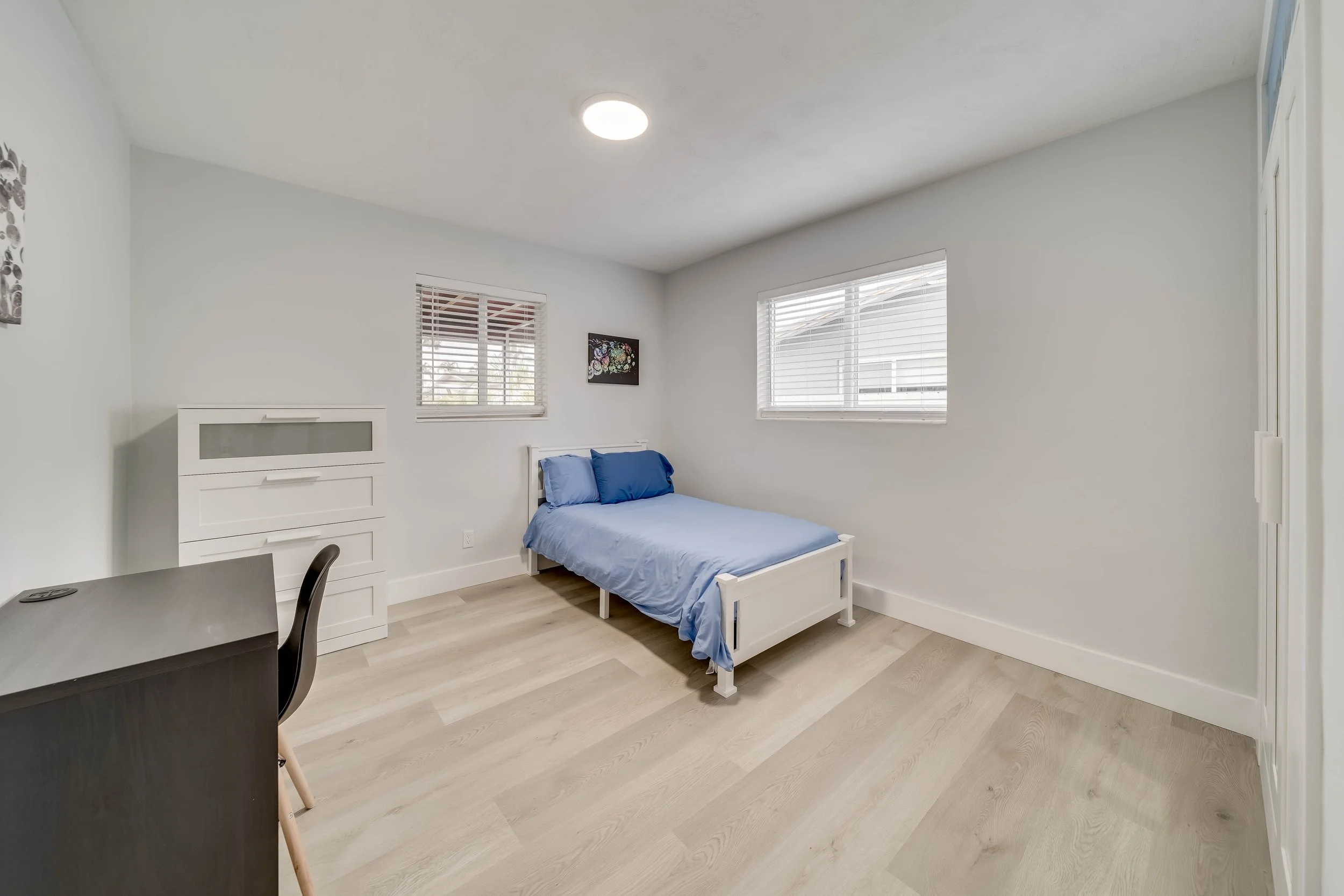 A minimalist bedroom with light-colored wooden floors, white walls, and two windows with blinds. There is a small white bed with blue bedding in the corner, a white dresser, a black chair, and framed artwork on the wall.