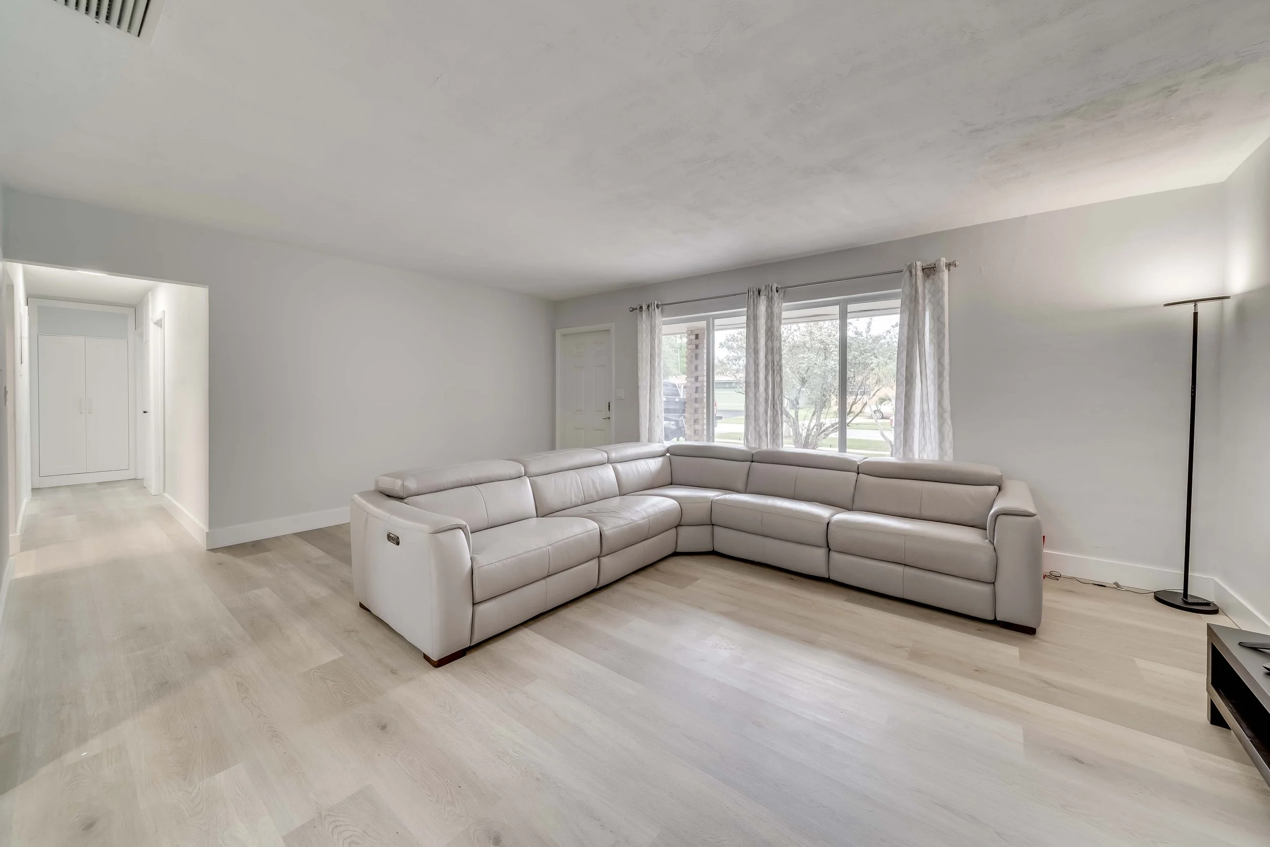 A living room with a white leather sectional sofa, large window with curtains, a floor lamp, and light wood flooring.