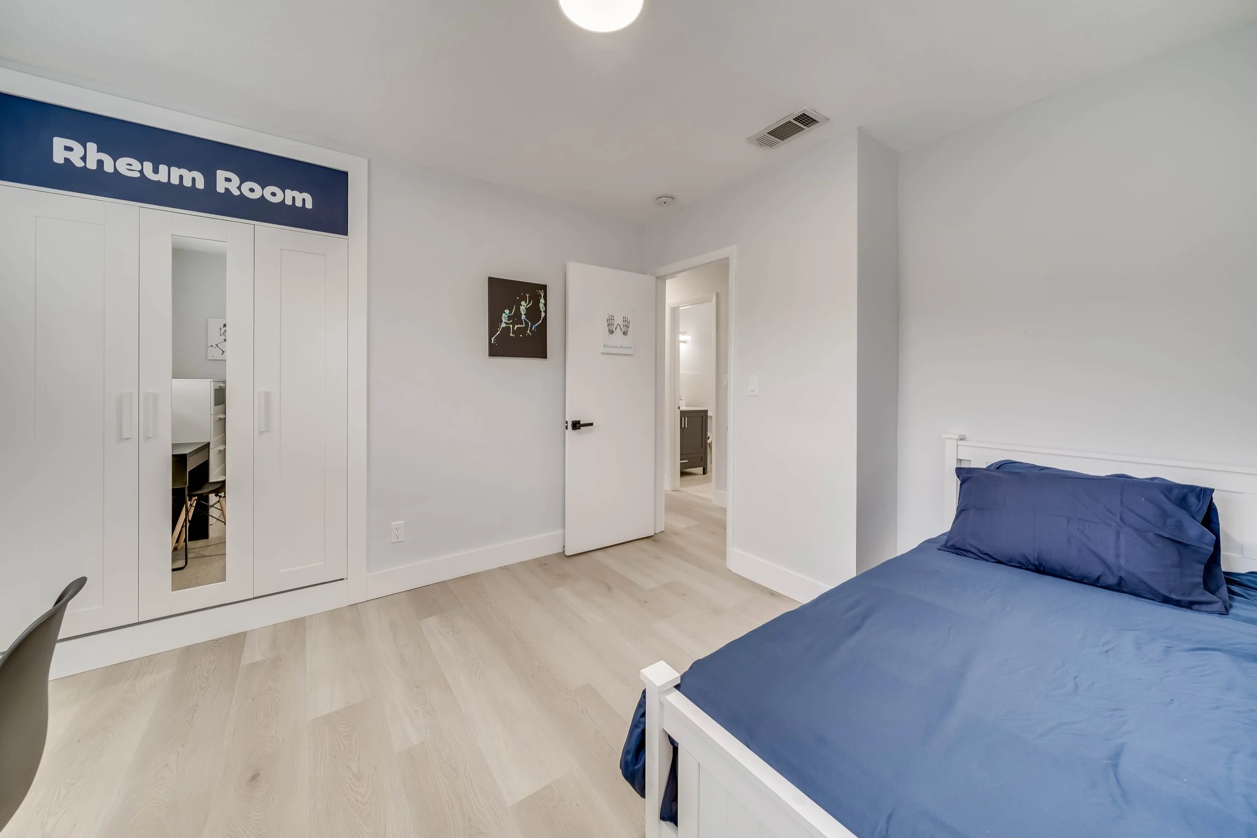 A clean, modern bedroom with a white bed frame and navy blue bedding, a white wardrobe with mirrored doors, and a small doorway leading to an en-suite bathroom. The walls are painted white, and there are some framed artworks hanging on the wall.