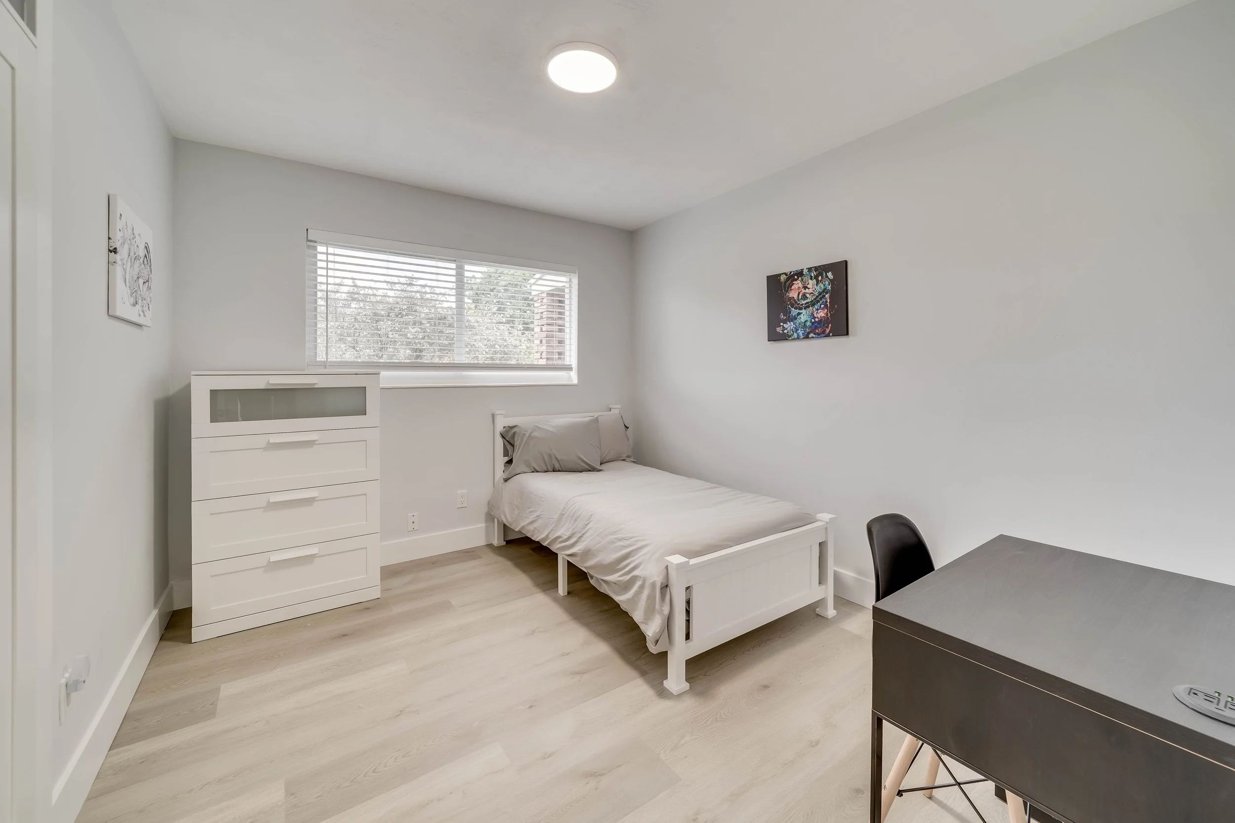 A minimalist bedroom with white walls, a single bed with gray bedding, a white dresser, a small dark table, and abstract art on the walls. There is a window with blinds letting in natural light.