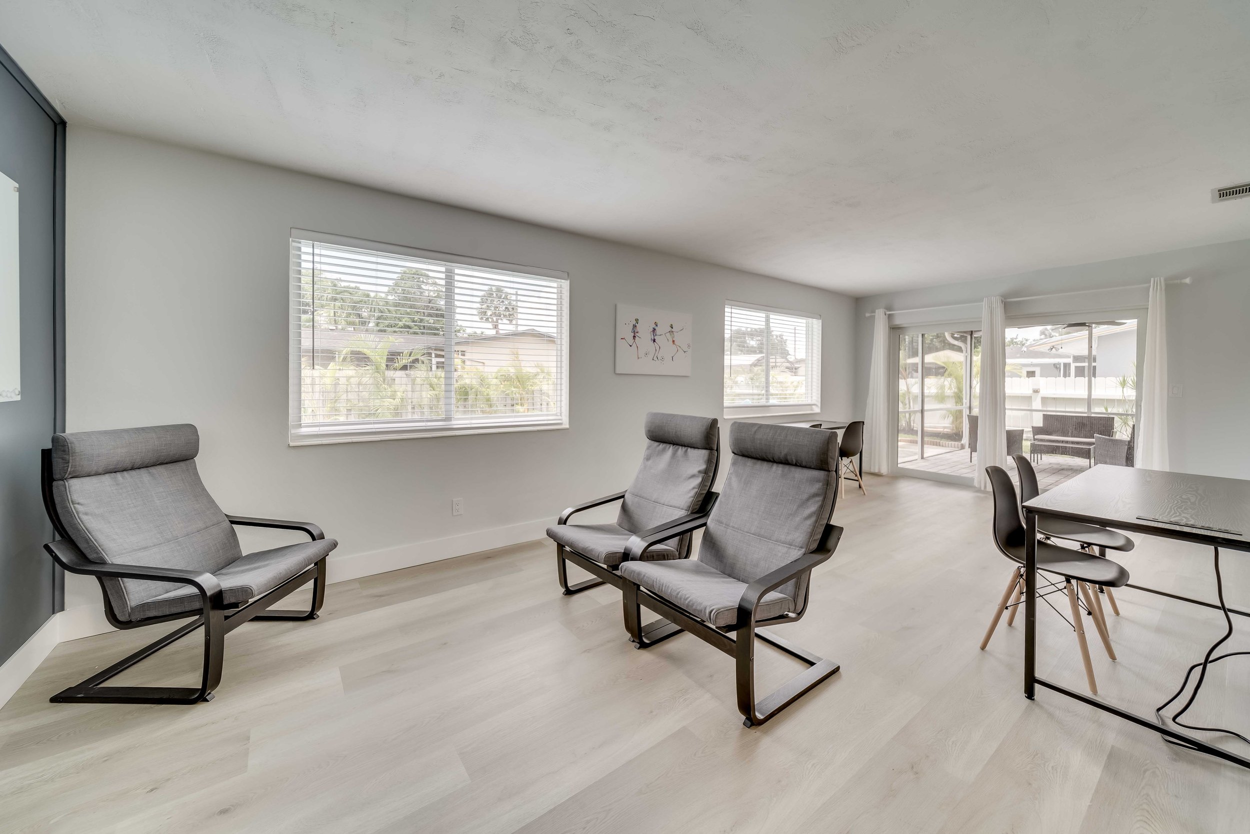 Living room with large windows, gray chairs, white walls, and a sliding glass door leading to an outdoor patio.