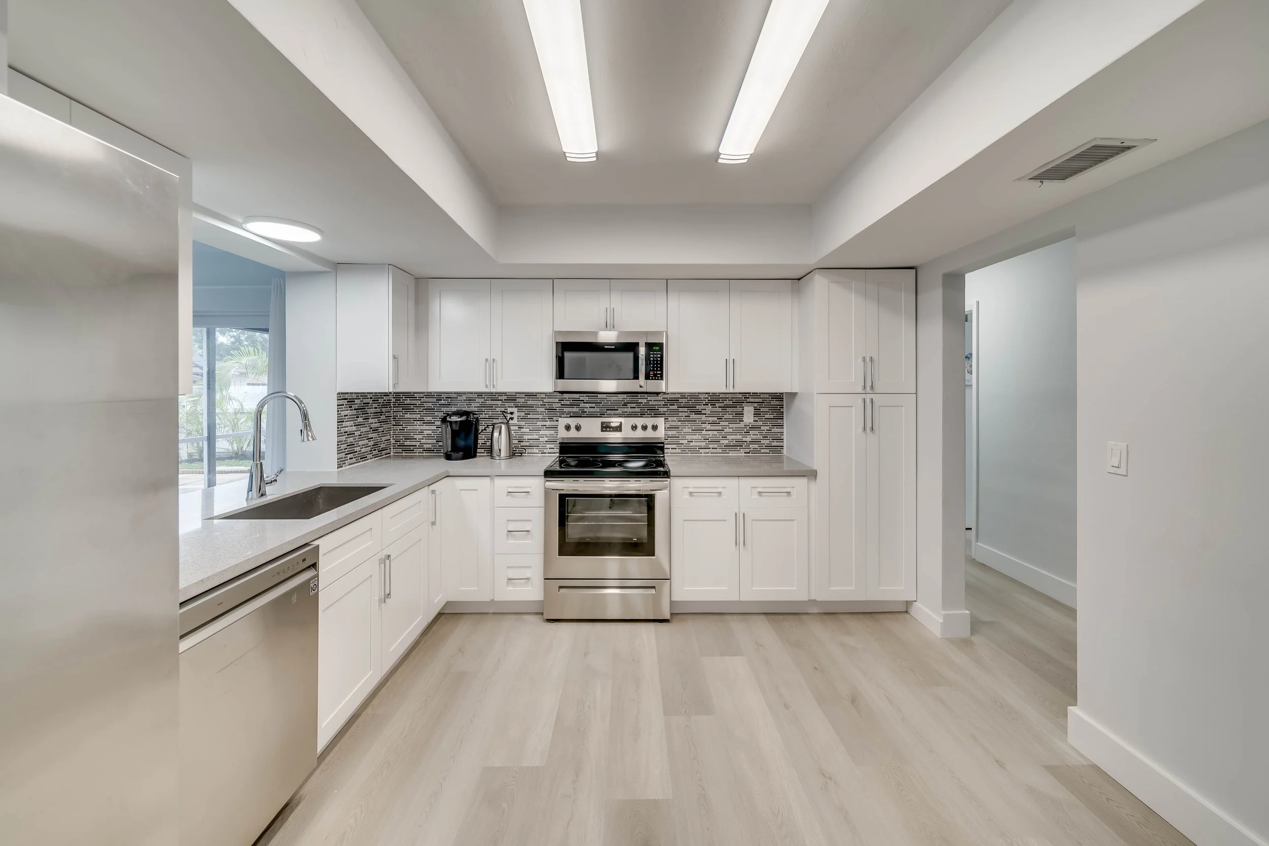 Modern kitchen with white cabinets, stainless steel appliances, gray mosaic tile backsplash, a sink with a faucet, and light wood flooring.