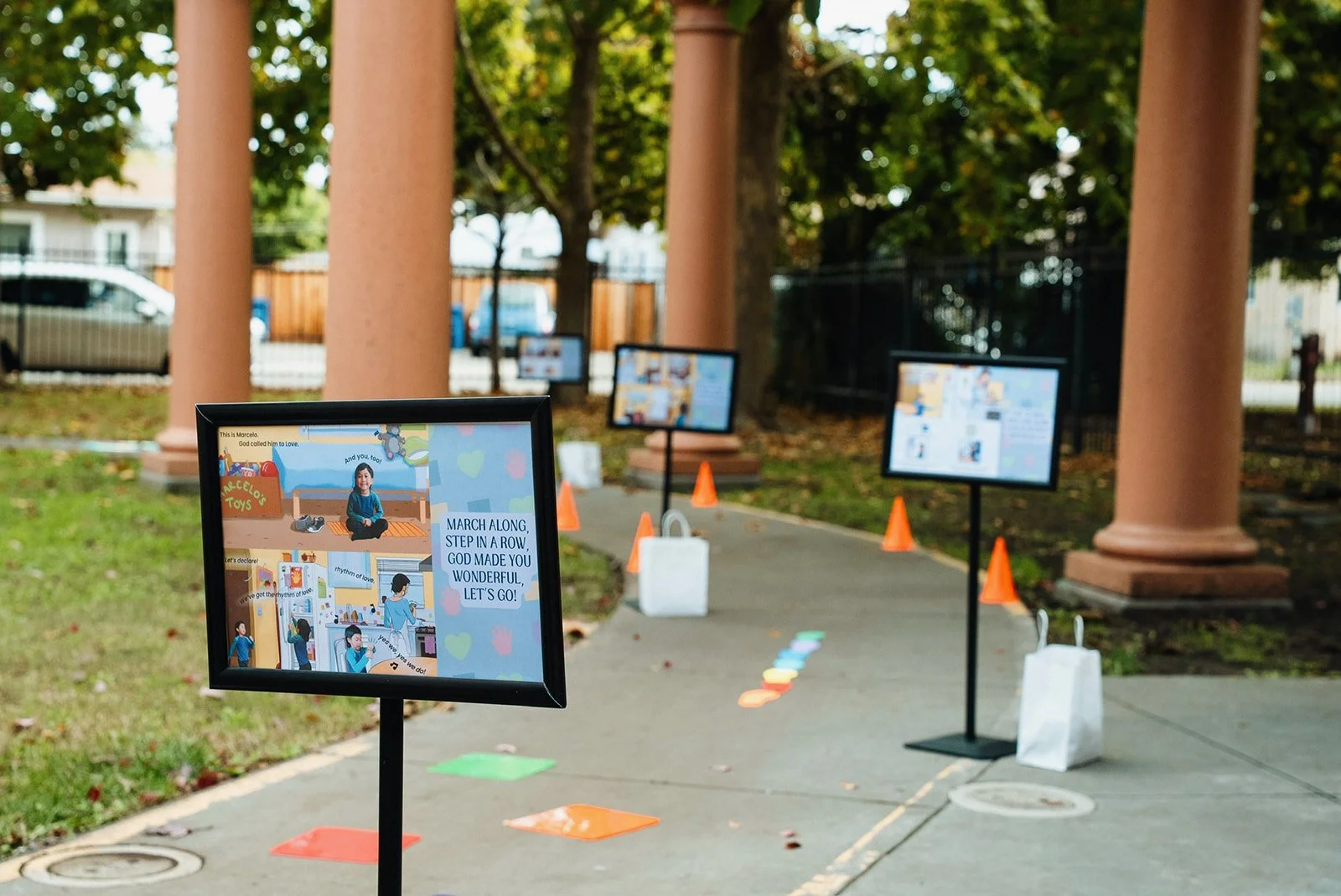 Outdoor display with animated comic panels on black frames, set along a sidewalk with orange cones and small white bags, surrounded by trees and pillars.