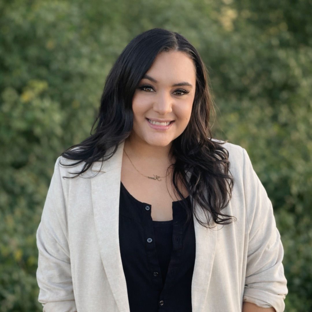 A smiling young woman with black wavy hair wearing a black top and a light-colored blazer, standing outdoors in front of green trees.