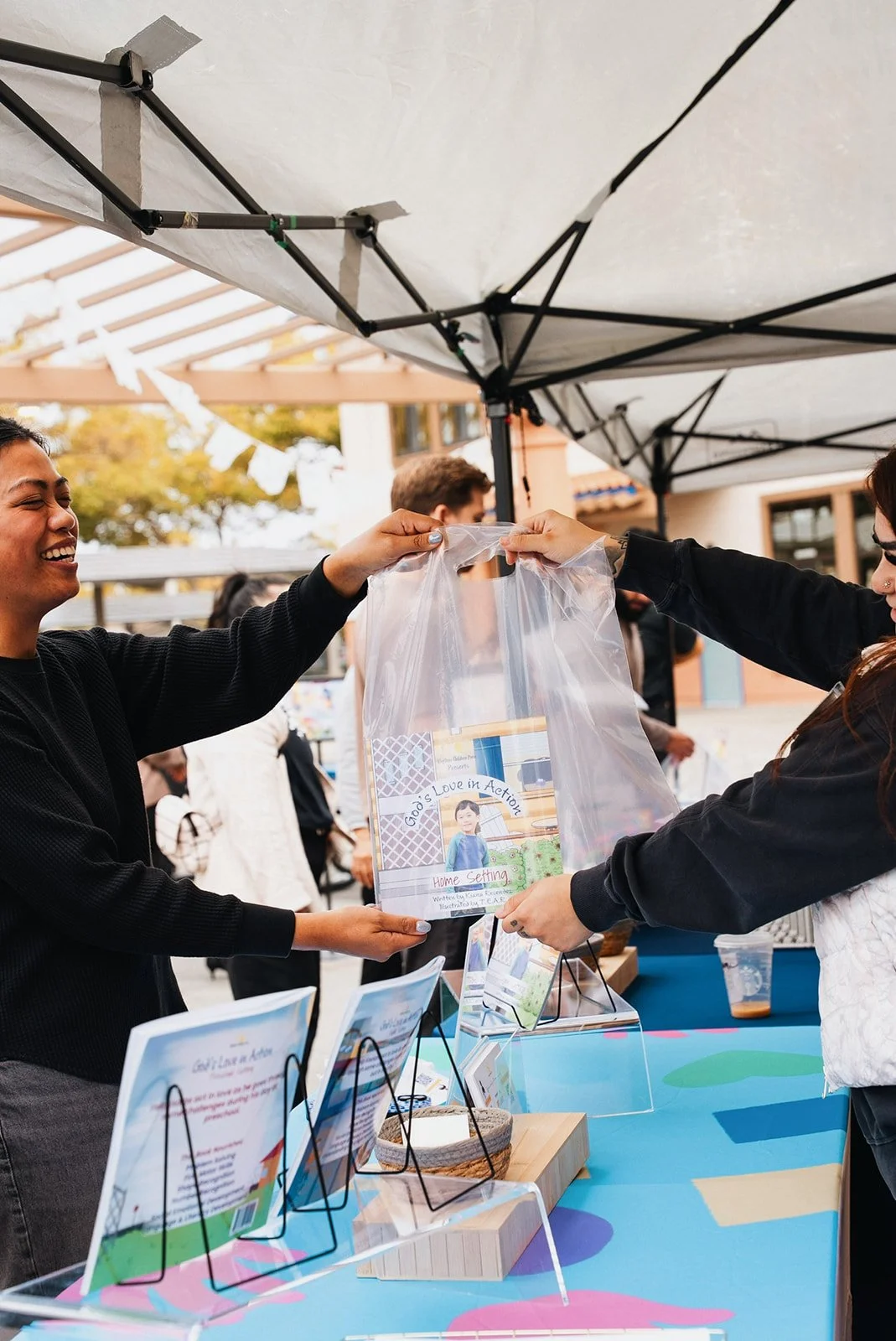 Two women at an outdoor booth exchanging a clear plastic bag with a book inside, surrounded by informational signs, under a canopy, with people in the background.
