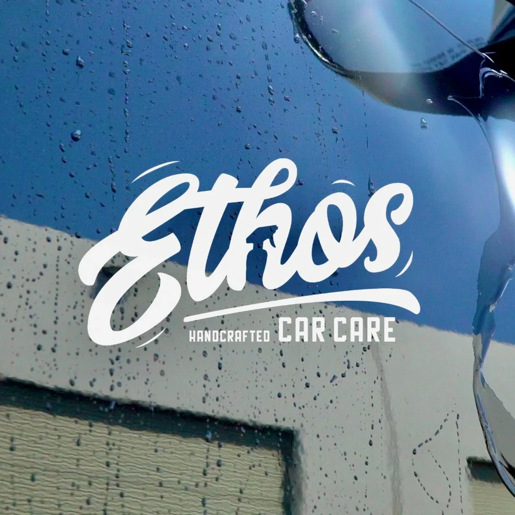 A car window with water droplets and a reflection of the sky. White text overlay reads 'Estros Handcrafted Car Care'.