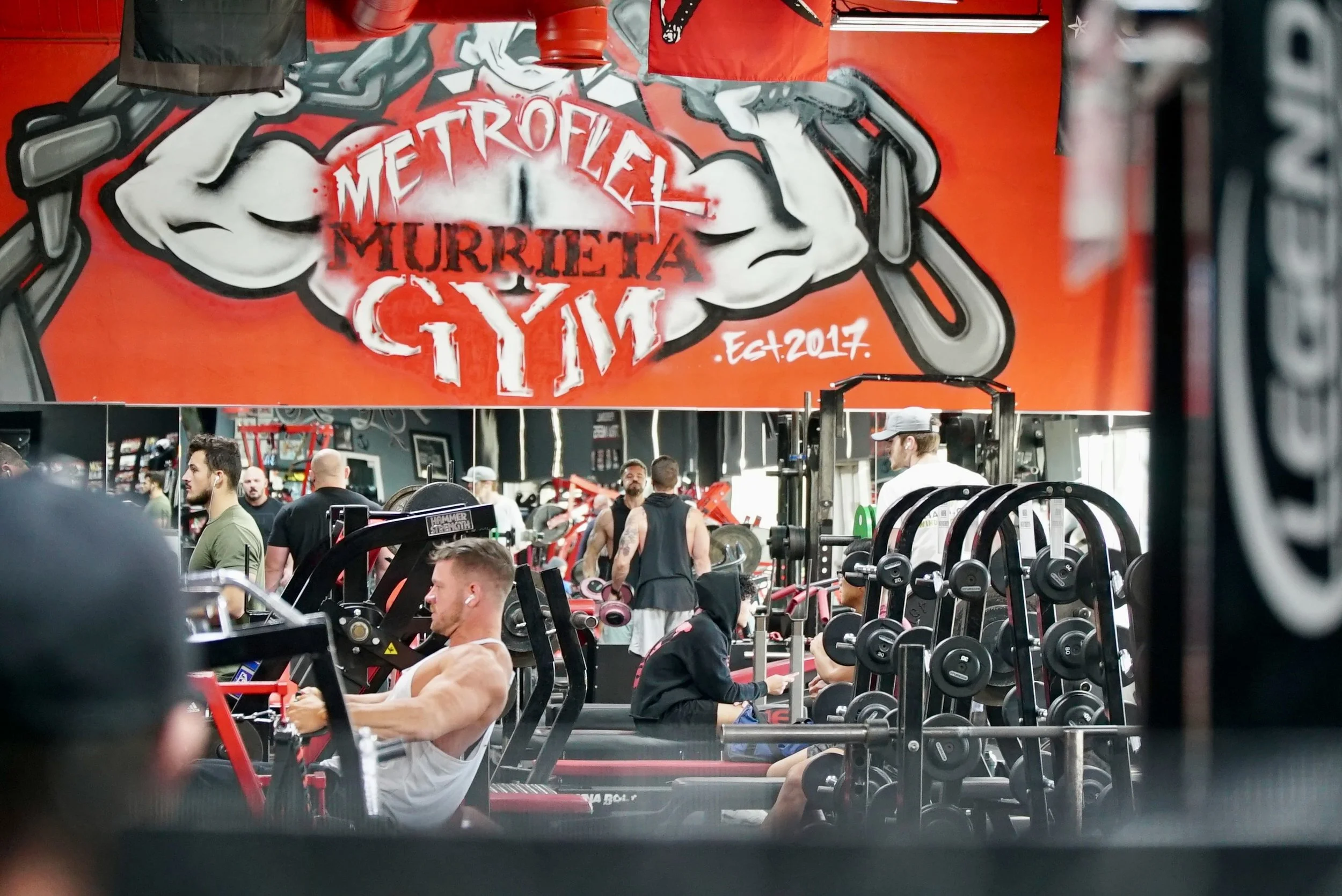 People working out at a gym with a large Metrolife Murrieta gym logo banner hanging above. The gym has various exercise equipment and weights, with some individuals in workout attire engaged in different physical exercises.