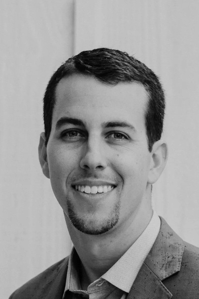 Black and white portrait of a young man with short dark hair, light skin, wearing a suit jacket and button-up shirt, smiling, with a plain light-colored background.